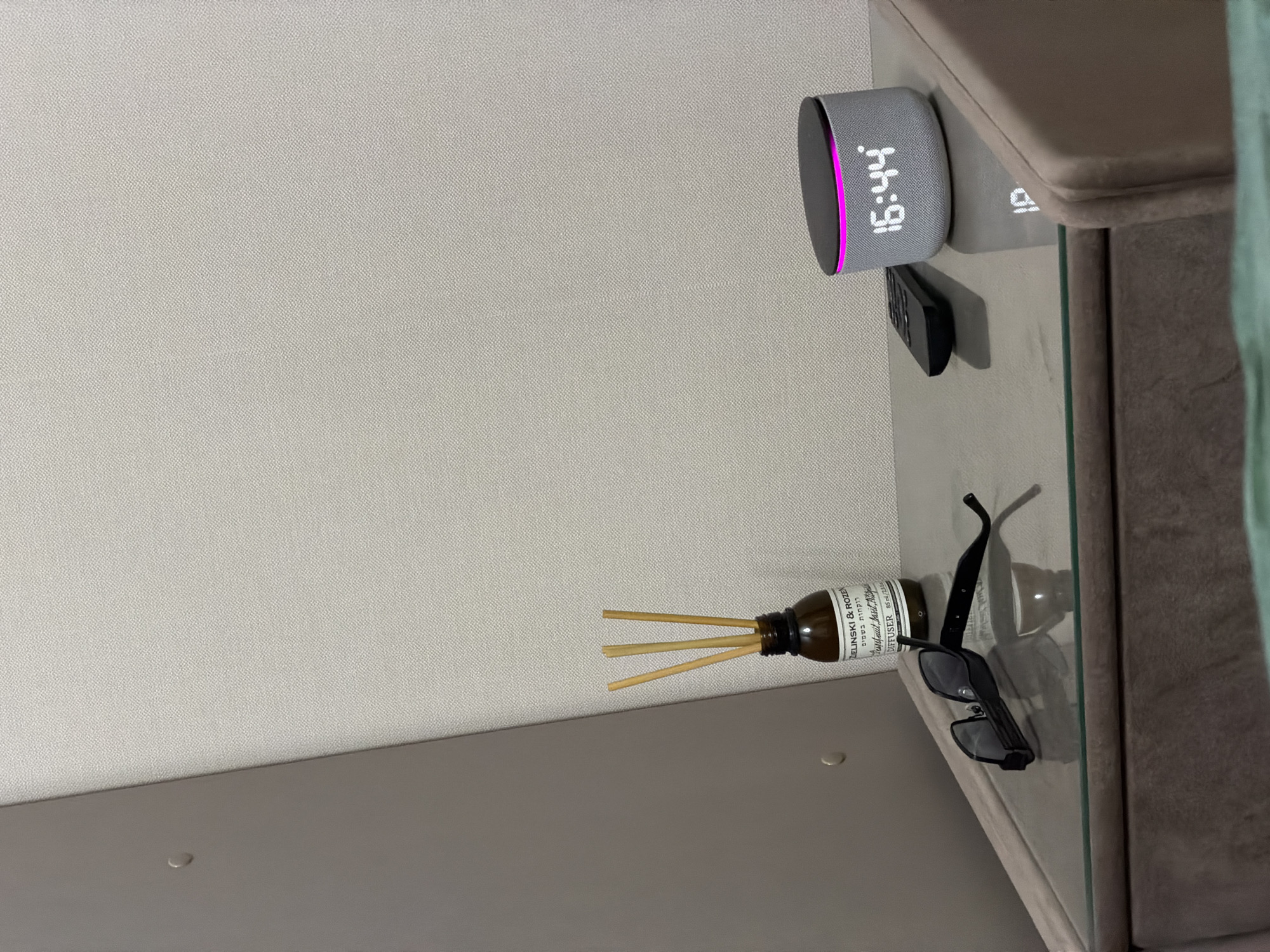 A bedside table in a room likely in Kazan, Russia, holds several items. On the glass surface of the table, a pair of black sunglasses is placed. To the right of the sunglasses is a small, dark remote control. Next to the remote is a cylindrical smart speaker with a pink illuminated ring around its top edge, displaying a digital time of "16:44". Behind the sunglasses, a brown glass bottle with a label, containing three reeds, likely a reed diffuser, stands upright. The background consists of a textured light-colored wall and a darker, angled surface on the left. A plush, neutral-colored chair or sofa is partially visible on the right. The overall scene depicts a quiet, uncluttered space.