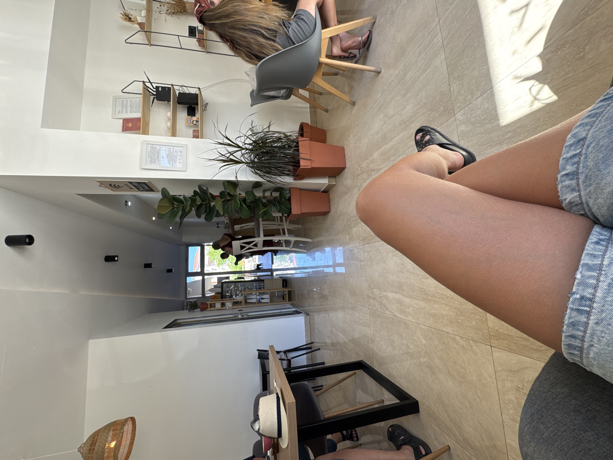 The image captures an indoor scene, likely a cafe or restaurant in Budva, Montenegro. The foreground features the legs of a person wearing denim shorts and black slides, with their legs crossed. In the mid-ground, a wooden table with a straw hat and some dishes is visible, accompanied by chairs. Further into the cafe, more seating areas with chairs and tables are present. Several potted plants, including a large ficus, add greenery to the space. Shelving units display various items, including books and decorative objects. In the background, through a doorway, a glimpse of the exterior shows trees and buildings, suggesting an urban environment. The flooring is tiled with a beige, marble-like pattern. The lighting appears to be natural, supplemented by artificial ceiling lights and a pendant lamp. The overall atmosphere seems relaxed and casual.