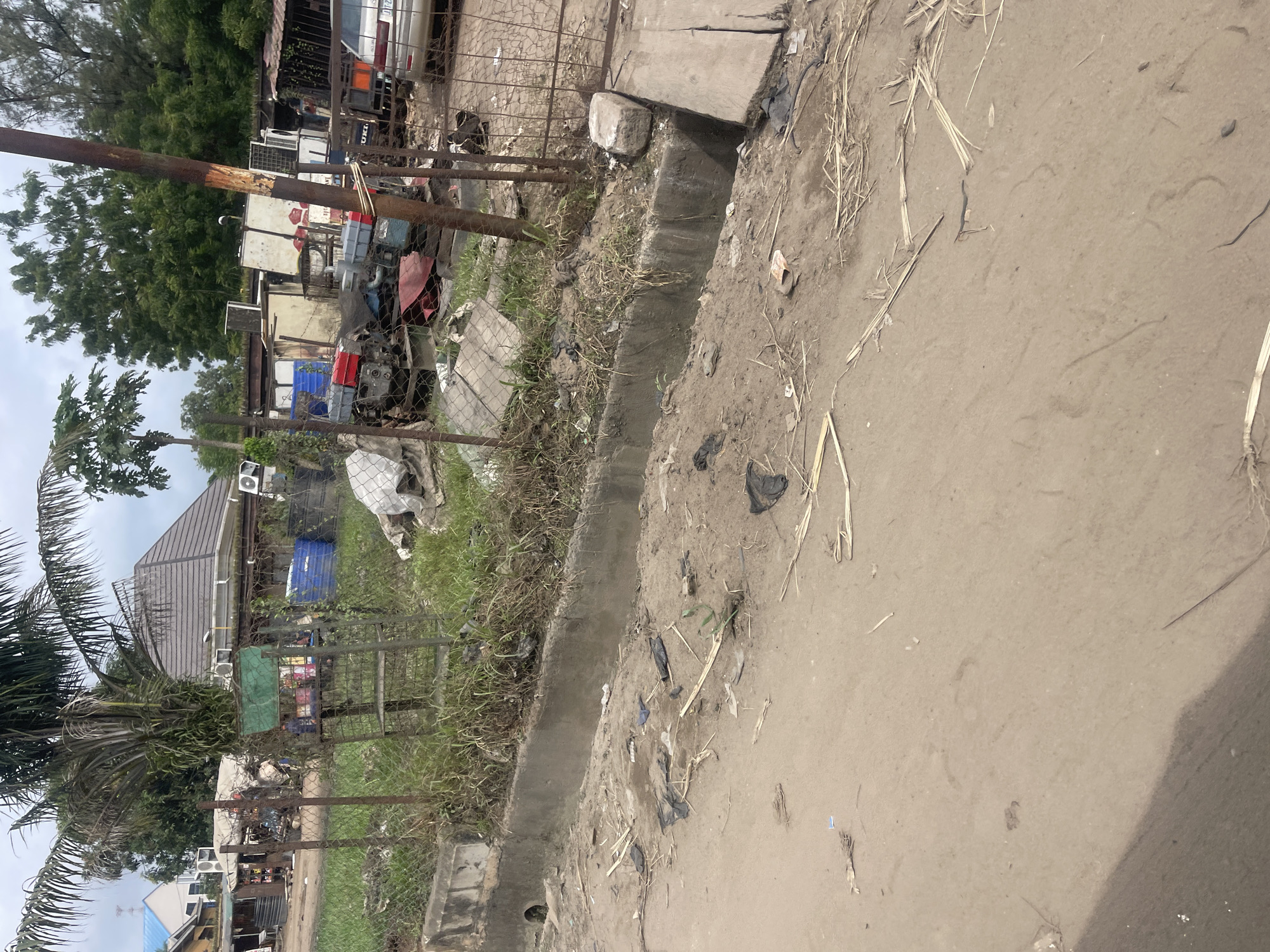 This image depicts a cluttered outdoor scene in Onitsha, Nigeria. In the foreground, a sandy, unpaved surface is visible, littered with dried grass, straw, and black plastic bags. A concrete drainage channel runs along the edge of the scene, also containing debris. Beyond the drainage channel, a chain-link fence encloses an area filled with various objects. These include rusted metal shelves holding colorful items, large blue plastic barrels, scrap metal, what appears to be engine parts, and several white rectangular units resembling air conditioning units or generators. A palm tree stands prominently to the left, with its fronds reaching towards a cloudy sky. In the background, there are buildings with grey tiled roofs and smaller structures that seem to be shops or stalls. A silver car is parked within the fenced area, partially visible. The overall impression is one of an unkempt storage or workshop area, with a mix of everyday items and industrial equipment.