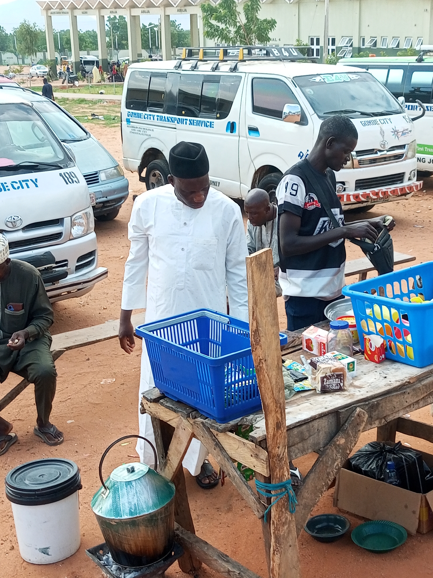 In an outdoor setting, likely a bus station or market, two men are visible. One man, wearing a white robe and a black cap, stands and appears to be interacting with items on a wooden table. On the table are several items, including two blue plastic baskets, boxes of food, a glass jar, and a loaf of bread. A large green metal pot sits on a charcoal brazier to the left of the table. To the right of the table is a cardboard box containing a black trash bag and a plastic water bottle. The second man, wearing a black and white striped shirt with the number "19" on it, stands and is observed putting items into a black bag. Another man, partially visible and wearing a green garment, is seated to the left. In the background, several white vans with "GOMBE CITY TRANSPORT SERVICE" branding are parked. The ground is covered in reddish-brown dirt. The environment suggests a commercial area with transportation services.