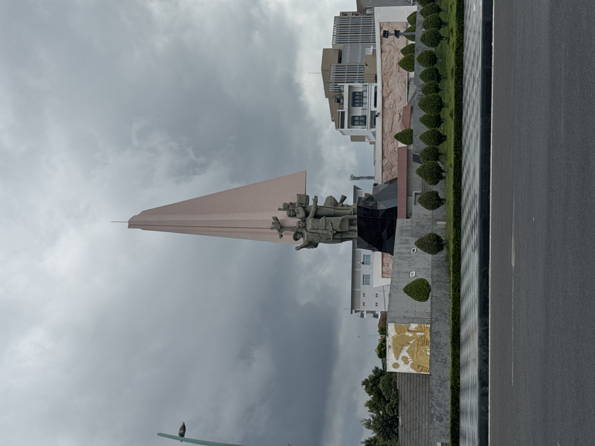 The image captures the Memorial of Heroes and Martyrs in Phan Thiết, Vietnam, under a heavily overcast sky. The central focus is a large, abstract monument with a tall, triangular, pale pink spire. At the base of the spire, a group of three stone figures is depicted: two adults holding a child, representing a family unit. To the left of the monument, a streetlamp with a teal pole and a white lamp head is visible.
Behind the monument, several buildings are present, ranging from low-rise structures to taller, modern buildings with distinct architectural features, including balconies and vertical lines of windows. The foreground of the scene shows a paved area with a checkered pattern, bordered by a manicured lawn with several evenly spaced, rounded evergreen bushes. Beyond the bushes, there is a low wall with a gold-colored relief depicting abstract shapes and figures. A paved road occupies the bottom portion of the image, showing lane markings. The overall scene suggests a public space, likely a plaza or roundabout, in a city environment.