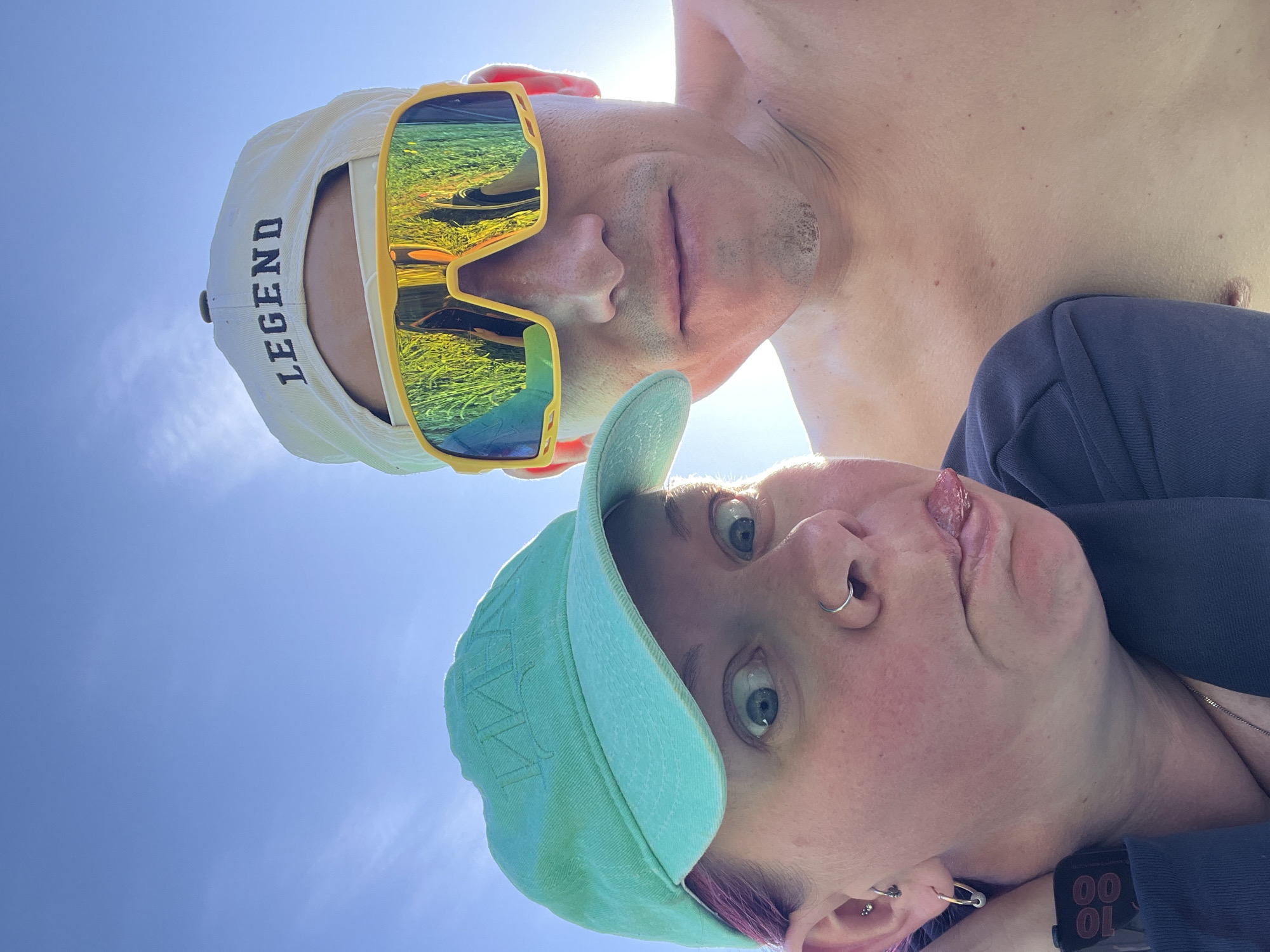 Two individuals are captured in a selfie-style, upward-angled photograph against a clear blue sky. The person at the top, a light-skinned man, wears a white baseball cap with the word "LEGEND" embroidered in black, along with yellow-framed sunglasses that reflect a landscape. The man appears shirtless. The second person, a light-skinned woman with purple-toned hair and multiple piercings, is positioned below. She is wearing a turquoise baseball cap.
It is not possible to identify the exact location in the city of Chaikovsky, Russia based on the given image.