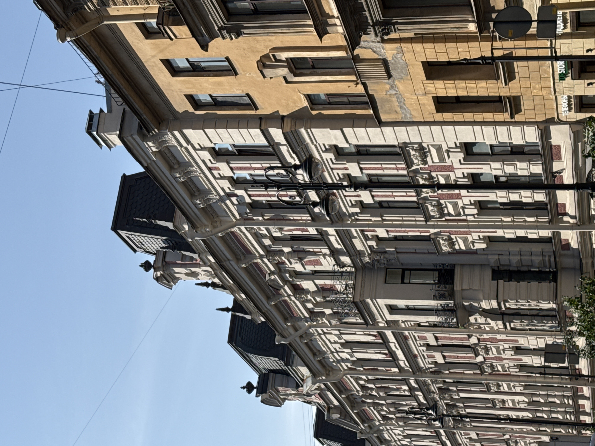 The image depicts a street view of historical buildings in Saint Petersburg, Russia, under a clear blue sky. The buildings exhibit classic St. Petersburg architecture with ornate facades, mansard roofs, and numerous windows. The building on the left is lighter in color, possibly cream or white, with red accents and intricate decorative elements. The building to its right is a warm yellow or ochre color, showing some signs of age and wear, with darker-framed windows. A street lamp with two globes stands in front of the lighter building. Overhead, thin wires are visible against the sky. In the foreground, a portion of a tree with green foliage is visible at the bottom. The overall impression is one of historical urban architecture bathed in sunlight.