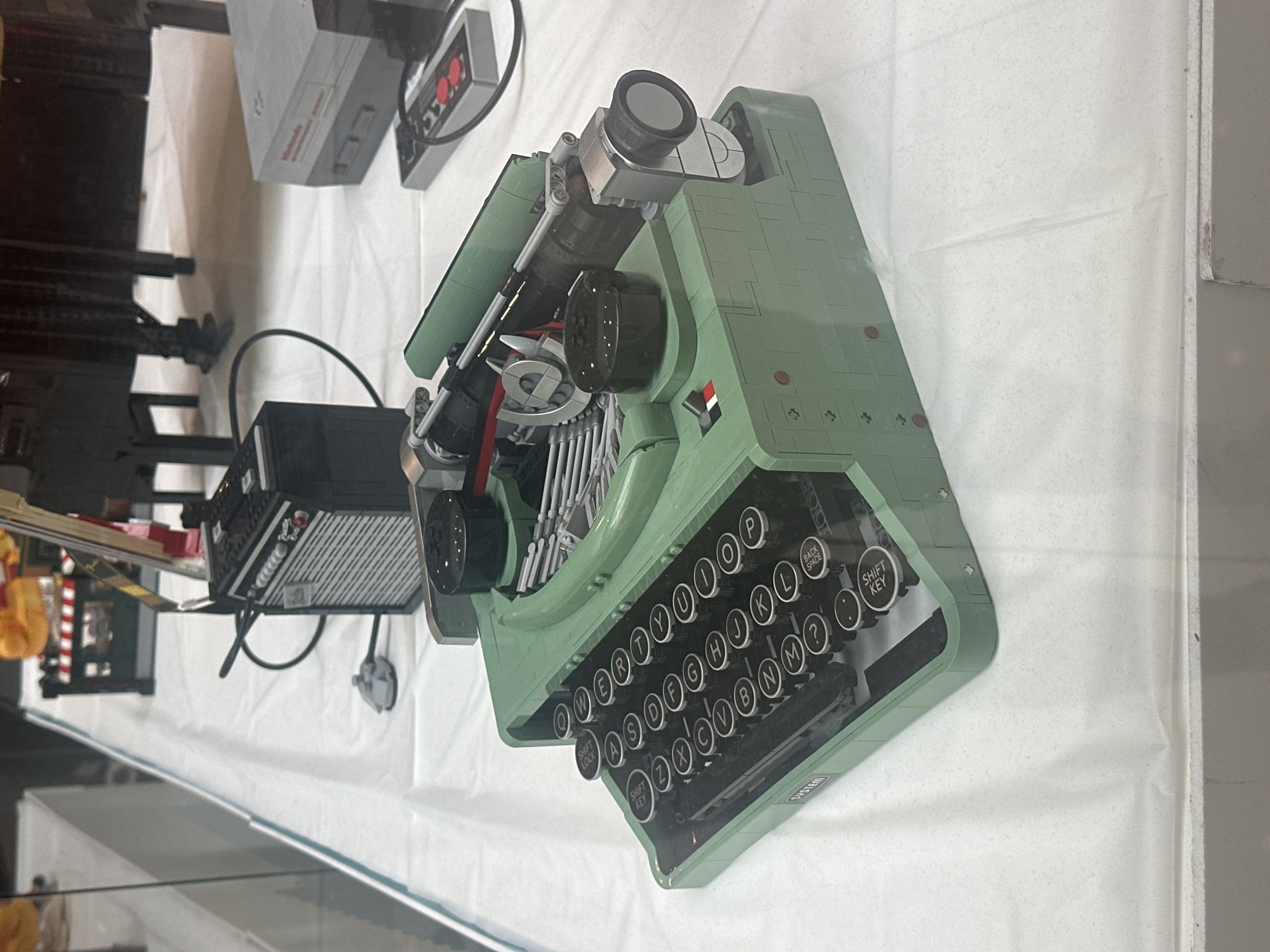 This media file captures a close-up, overhead view of a light green, LEGO-constructed replica of a vintage typewriter. The typewriter is the central and only prominent object in the frame, placed on a white, textured surface that appears to be a tablecloth or display cloth.
The typewriter is detailed, showcasing individual keys with letters and symbols, as well as mechanical elements like the carriage and typebars. Some keys are labeled with letters such as "Q", "W", "E", "R", "T", "Y", "U", "I", "O", "P", "A", "S", "D", "F", "G", "H", "J", "K", "L", "Z", "X", "C", "V", "B", "N", "M". There are also special function keys visible, including "SHIFT LOCK", "SHIFT KEY", and "BACK SPACE". A circular dial is present on the right side of the typewriter, and the top features what appears to be a magnified viewer or lens. A small red, white, and black emblem is affixed to the side of the typewriter body.
To the left of the typewriter, partially visible, is a black electronic device with a ridged surface, possibly a radio or audio equipment, also constructed from LEGO. Further in the background, a grey and red LEGO Nintendo Entertainment System (NES) console with its controller is visible. The overall scene suggests a display or exhibition of LEGO models. There is no indication of people, time of day, weather, or specific location cues beyond the general context of Sofia, Bulgaria, provided separately. The mood conveyed is one of meticulous craftsmanship and nostalgia. The text "SYSTEM" is visible on a small plate on the front of the typewriter.