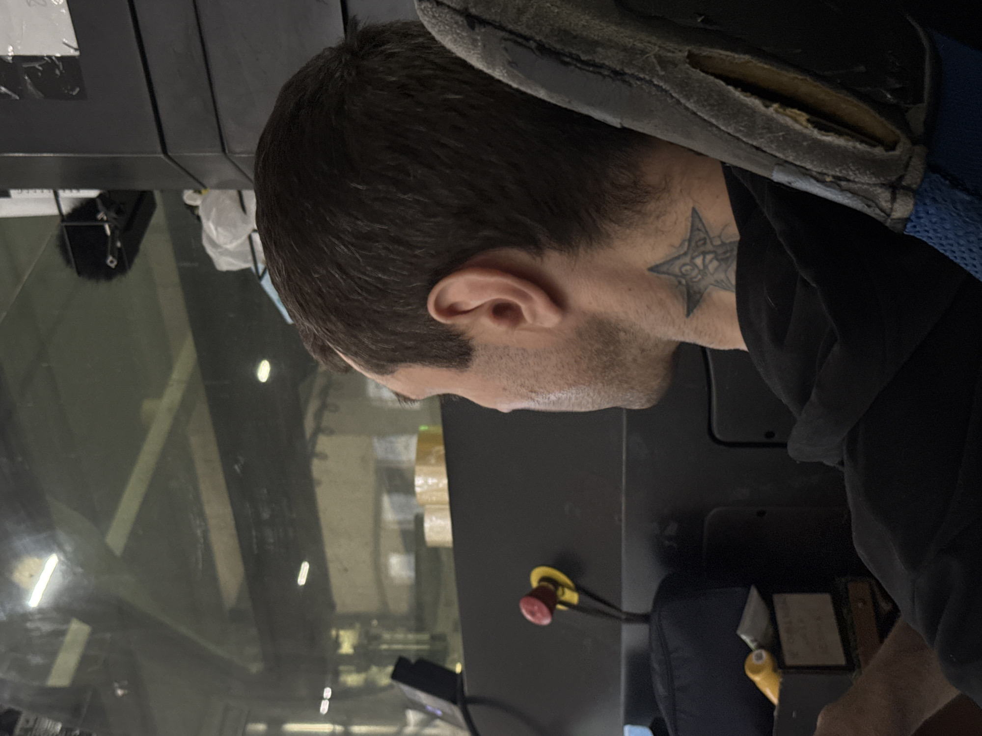 A man with dark hair and a star tattoo on his neck is seated. He is wearing a dark shirt. The background features machinery visible through a window, and controls with a yellow and red button are present. The location is suggested to be Perm, Russia.