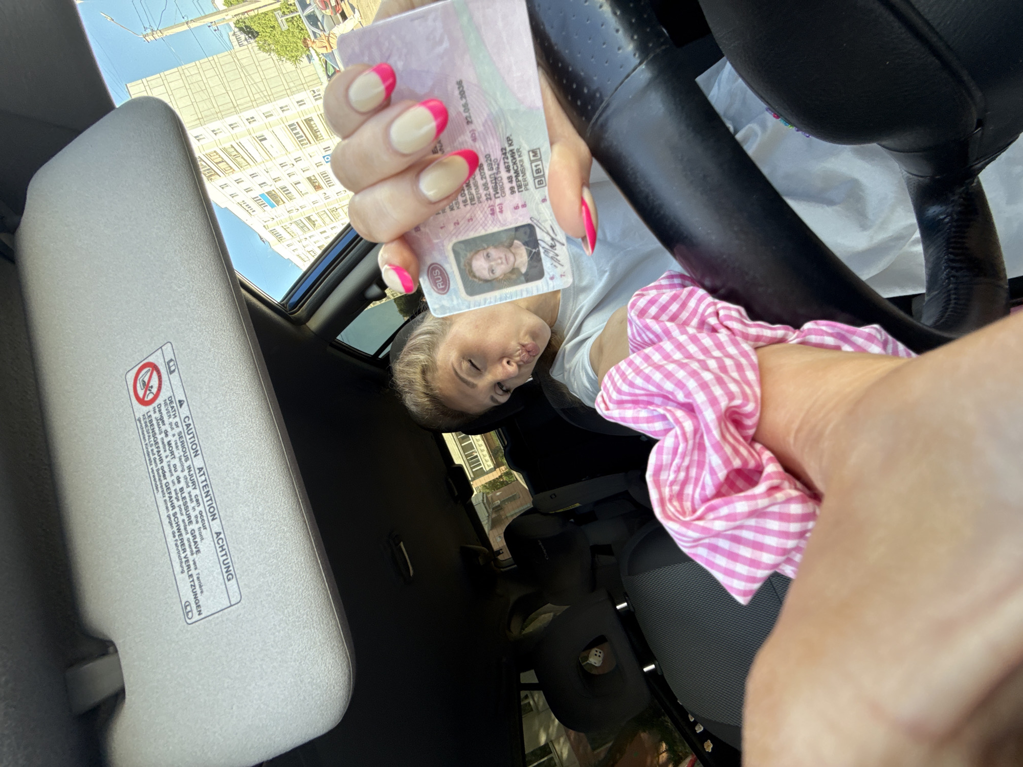 The image, taken from inside a car, shows a woman holding up what appears to be a Russian driver's license. The license has a photo of the woman and contains the acronym RUS. The woman has blond hair, fair skin, and is wearing a white shirt. She has pink and white nail polish. The image also shows a black steering wheel, a pink and white checkered scrunchie, and a sun visor with a warning label written in multiple languages. The buildings visible through the car's roof suggest a city setting. The driver's license shows the name of Permskii Kr, implying the city in question may be Chaikovsky, Russia.