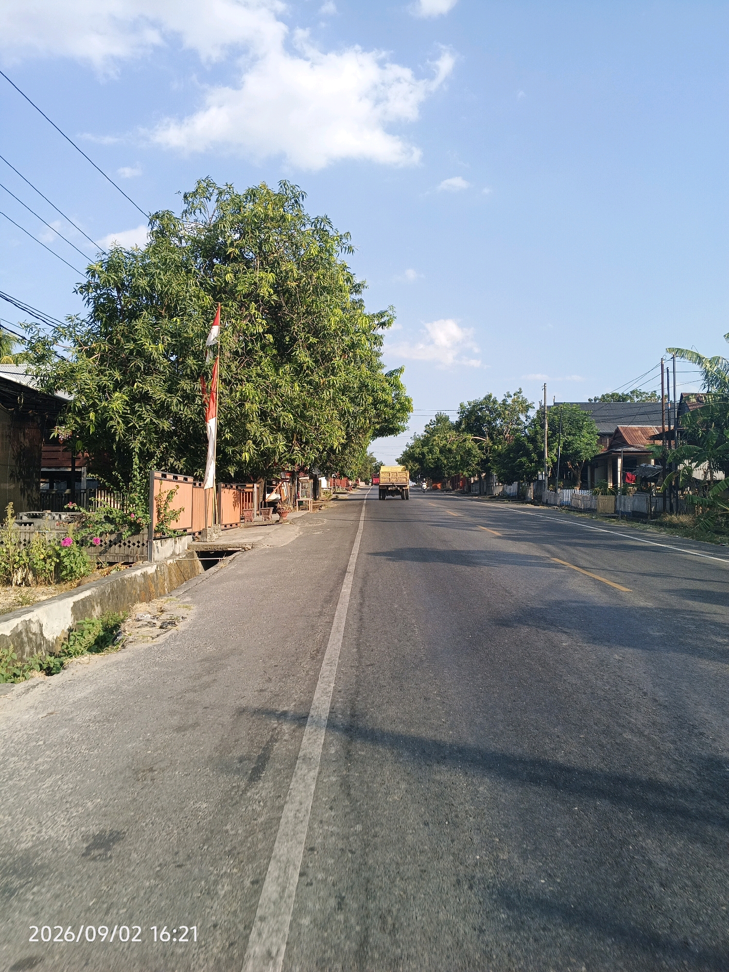 The image is an outdoor, eye-level, horizontal, full-shot photograph taken on a sunny day in Pinrang, Indonesia. It depicts a road with a yellow stripe down the middle, running through a residential area. The road is paved and has a white stripe on the left side. On the left side of the road, there is a sidewalk with a concrete drainage ditch, a low concrete wall, and vegetation including green plants and some pink flowers. Behind the sidewalk, there are houses and a large tree with green foliage. An Indonesian flag, red and white, is visible on a pole near the tree. On the right side of the road, there are more houses with various roof types, some with visible fences and trees. A yellow flatbed truck is driving away from the camera on the road in the distance. The sky is blue with scattered white clouds. The timestamp in the lower left corner reads "2026/09/02 16:21".