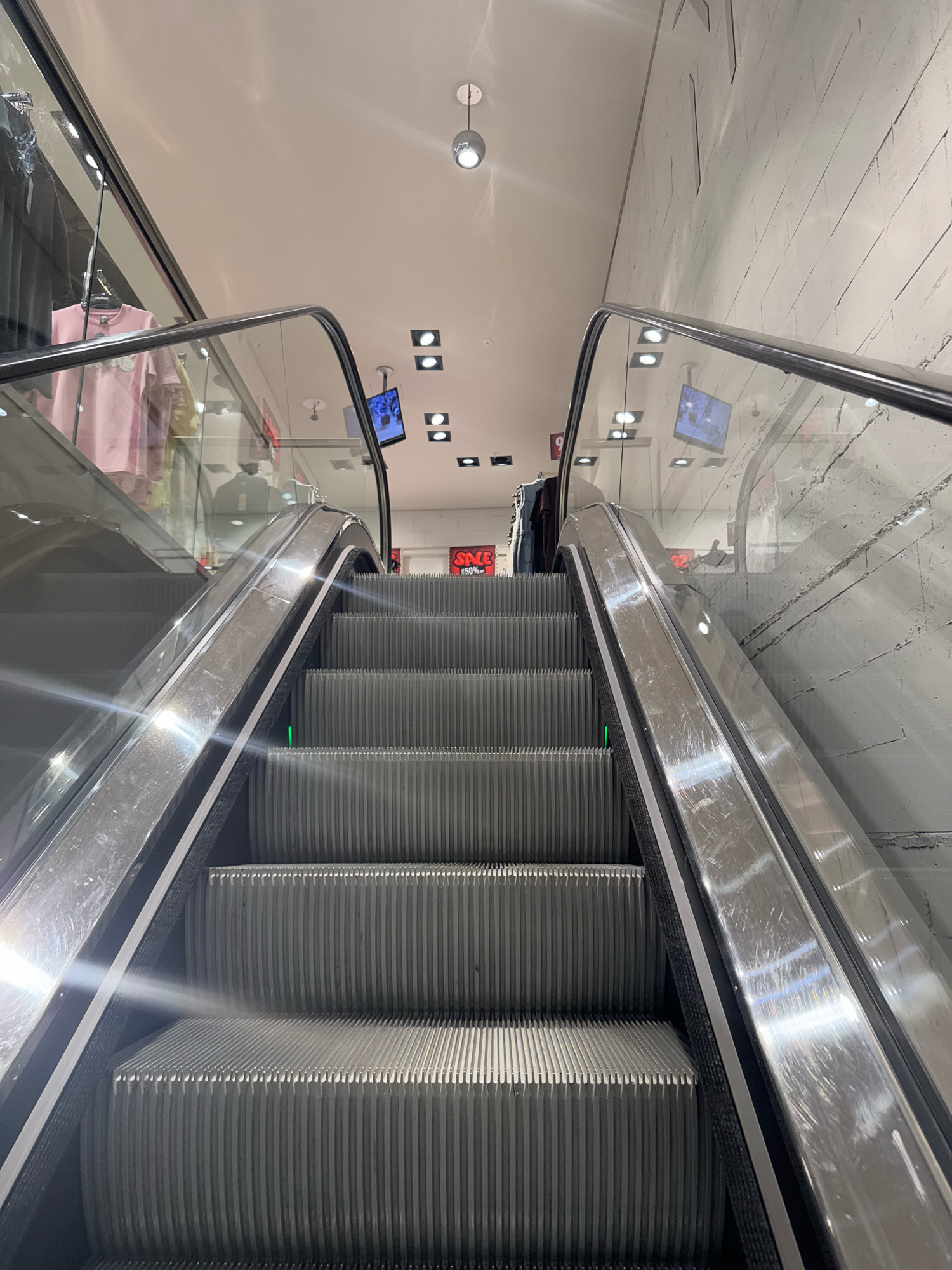 The image shows a close-up, low-angle view of an escalator in what appears to be a retail store. The escalator steps are made of metal with a ribbed texture. The handrails are also metal and appear to be slightly scratched. On the left side of the escalator, a glass barrier reveals clothing racks with shirts hanging, one of which is light pink. On the right side, a textured concrete wall with visible cracks and lines is present. Above the escalators, ceiling lights and screens displaying content are visible. There are no people clearly depicted in the image, though reflections in the glass suggest the presence of others. The scene is well-lit, with the lighting creating bright reflections on the metallic surfaces. The image does not provide any specific information to identify its location within the city of Nijmegen, Netherlands.