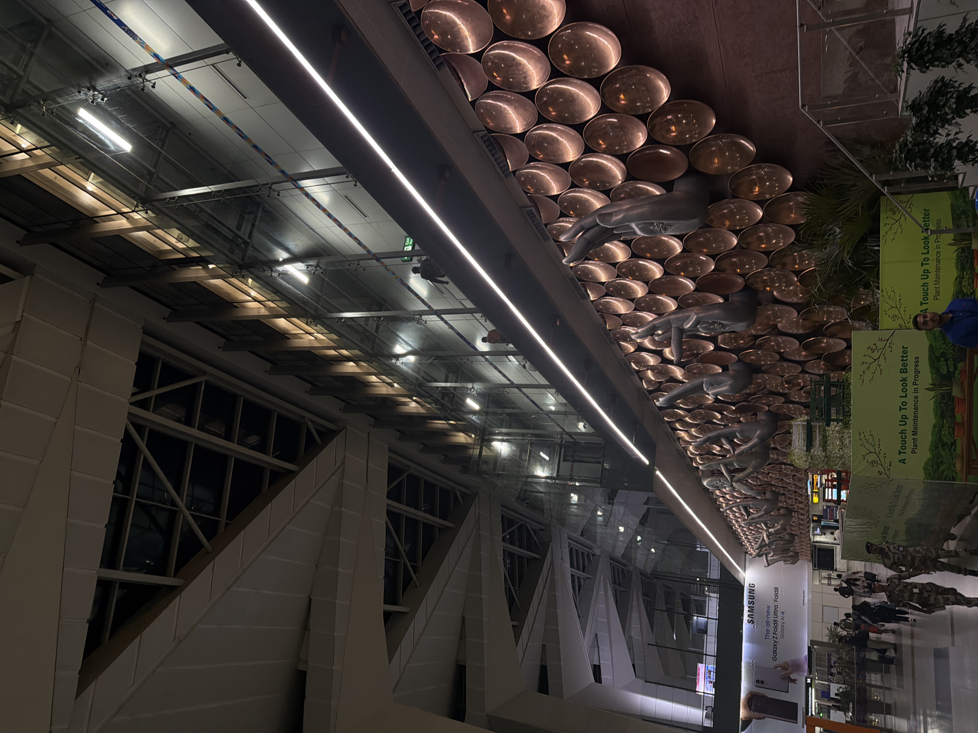 This image captures the interior of Terminal 3 at Indira Gandhi International Airport in Delhi, India. The scene is illuminated by artificial lights, highlighting the modern architecture and artistic installations. Dominating the frame is a large, ornate wall sculpture composed of numerous circular, copper-colored elements, interspersed with metallic hand sculptures in various mudras.
In the foreground, several people are visible. Two individuals in camouflage uniforms, presumably security personnel, are walking towards the left of the frame. Other pedestrians are also present, some walking, some standing, in what appears to be a concourse area.
A large Samsung advertisement for the Galaxy Z Fold8 Ultra is prominently displayed on a wall to the left. Further into the background, glimpses of escalators, glass partitions, and structural elements of the airport ceiling are visible. The overall environment suggests a busy, public space within a large international airport. The lighting creates a dramatic contrast between the illuminated areas and the darker, shadowed portions of the architecture.