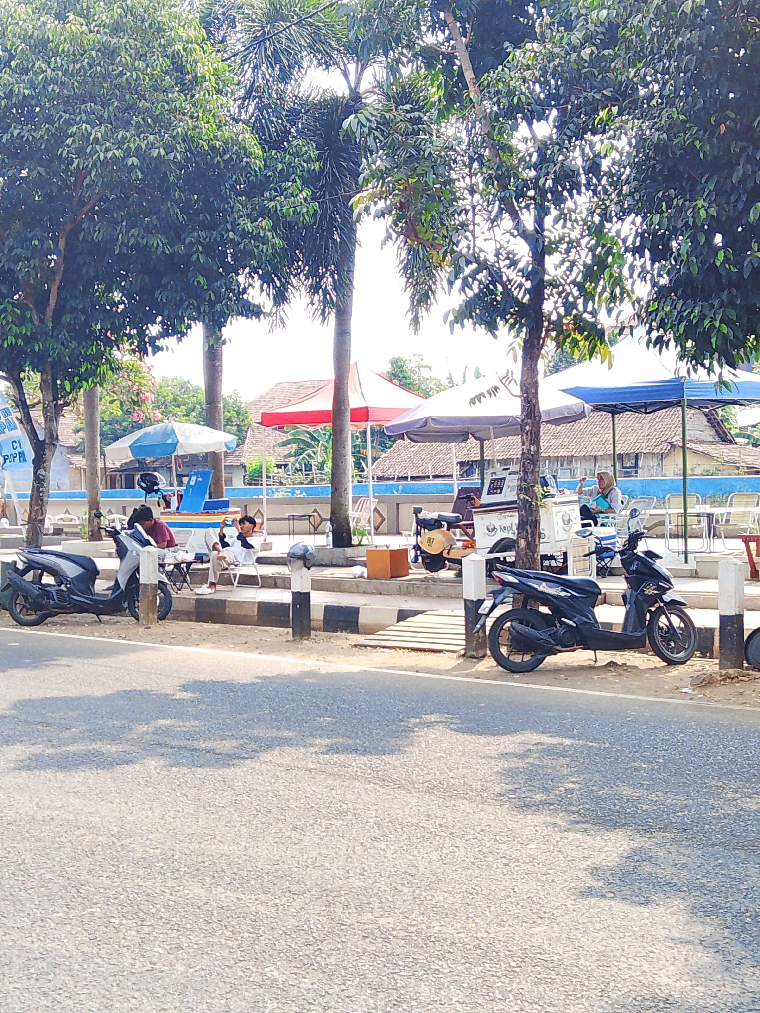 The image depicts a roadside scene in Potroyudan, Indonesia, on a sunny day. In the foreground, a paved road runs horizontally across the frame, with shadows of trees cast upon it. Behind the road, a sidewalk area is visible, separated by a curb with black and white striped markers.
Several parked motorcycles are present on the sidewalk. To the left, a silver scooter is parked. Further to the right, a black scooter is prominently displayed.
There are multiple people seated at small tables along the sidewalk. On the left, two young men are seated, one wearing a red shirt and the other a white shirt. They appear to be casually interacting. To their right, under a light-colored umbrella, a person wearing a headscarf is seated, holding a bag and looking towards the right.
A food or drink cart is situated behind the seated individuals, with a logo that includes "Kepl". Several umbrellas, including red, white, and blue ones, are set up to provide shade over seating areas. Beyond the sidewalk, a building with a tiled roof and a blue wall is visible. Trees with lush green foliage and palm trees are scattered throughout the scene, providing shade and contributing to the tropical atmosphere. The overall impression is of a relaxed, outdoor community gathering spot by the roadside.