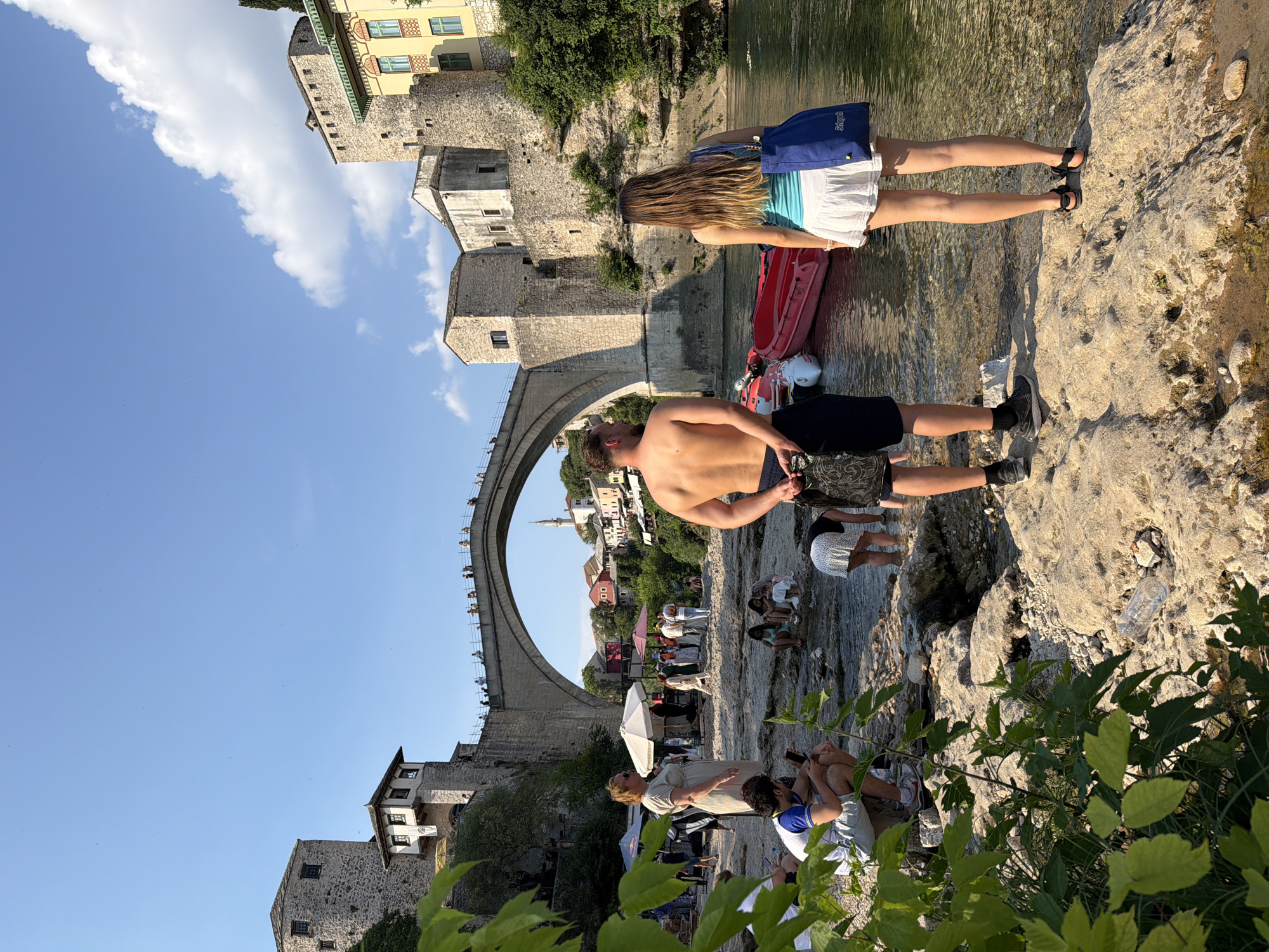 The image captures a sunny day by the Neretva River in Mostar, Bosnia and Herzegovina, with the iconic Stari Most (Old Bridge) dominating the background. Several people are present in the foreground and midground. In the immediate foreground, a man and a woman stand on a rocky riverbank, facing away from the camera. The man is shirtless, wearing dark shorts. The woman wears a light blue tank top and a white skirt, carrying a blue tote bag.
Further back, a group of people are gathered near the river's edge, some wading in the shallow water. One woman, in a light-colored dress, stands near an umbrella. The Stari Most, a UNESCO World Heritage site, arches gracefully over the river, with its stone towers and structure clearly visible. People can be seen walking on the bridge. Red kayaks or small boats are positioned in the river near the foreground subjects. The environment is characterized by the river, rocky terrain, and historic stone buildings that line the riverbanks. Green foliage is visible in the lower left corner of the frame. The sky is clear blue with a few scattered clouds.