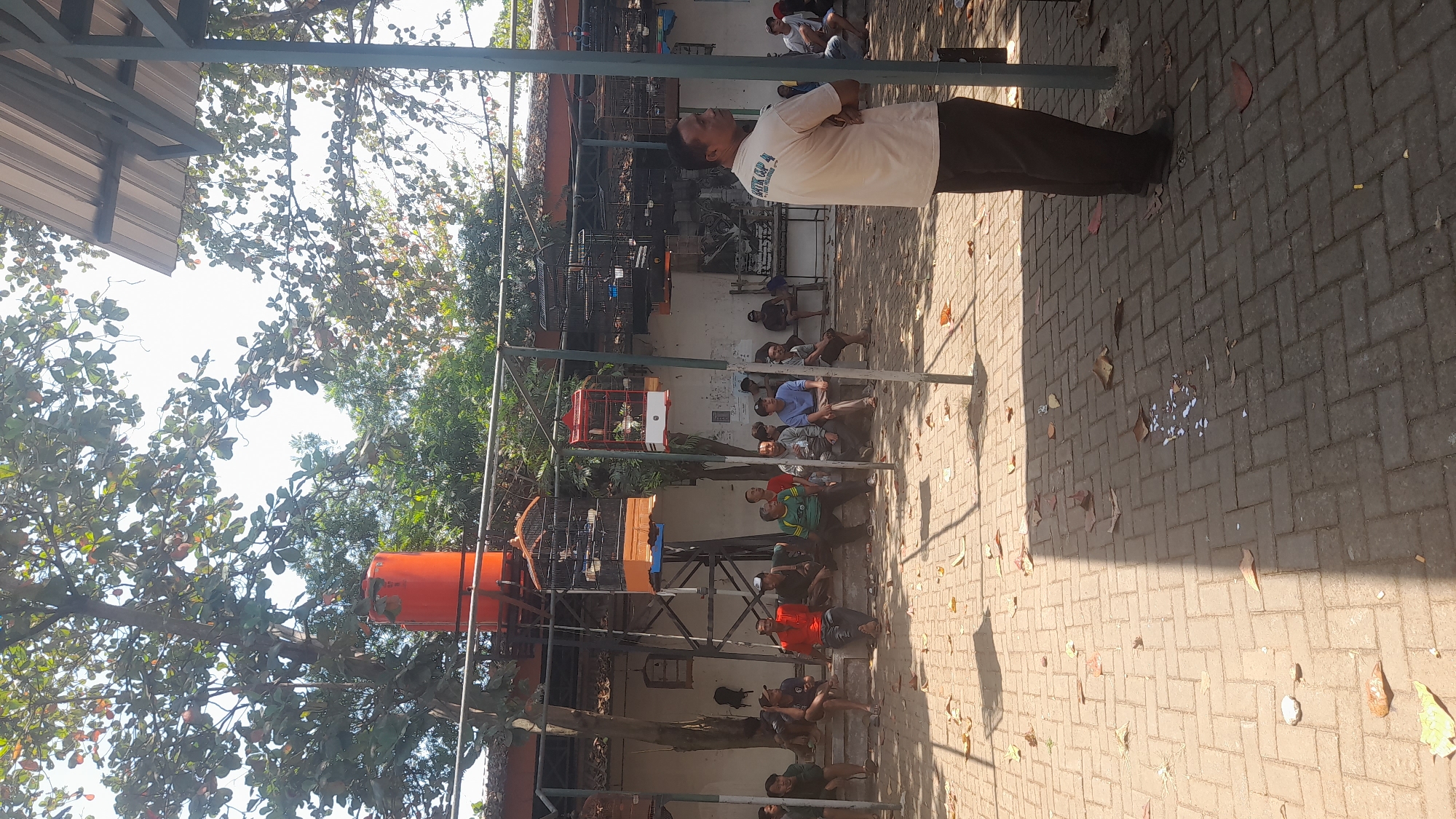 The image depicts an outdoor scene in Pekalongan, Indonesia, likely a bird competition venue. A group of men are seated on a raised concrete platform, observing something off-camera. In the foreground, a man in a beige t-shirt with text on the back and dark pants stands facing away from the viewer, arms crossed. The environment is paved with brick-like stones, scattered with fallen leaves. Several bird cages, some with birds inside, are suspended from metal frames, suggesting a bird-related event. A large orange water tank is visible in the background, supported by a metal structure. The scene is illuminated by sunlight, casting shadows on the ground. The overall atmosphere appears to be casual, with people gathered to watch or participate.