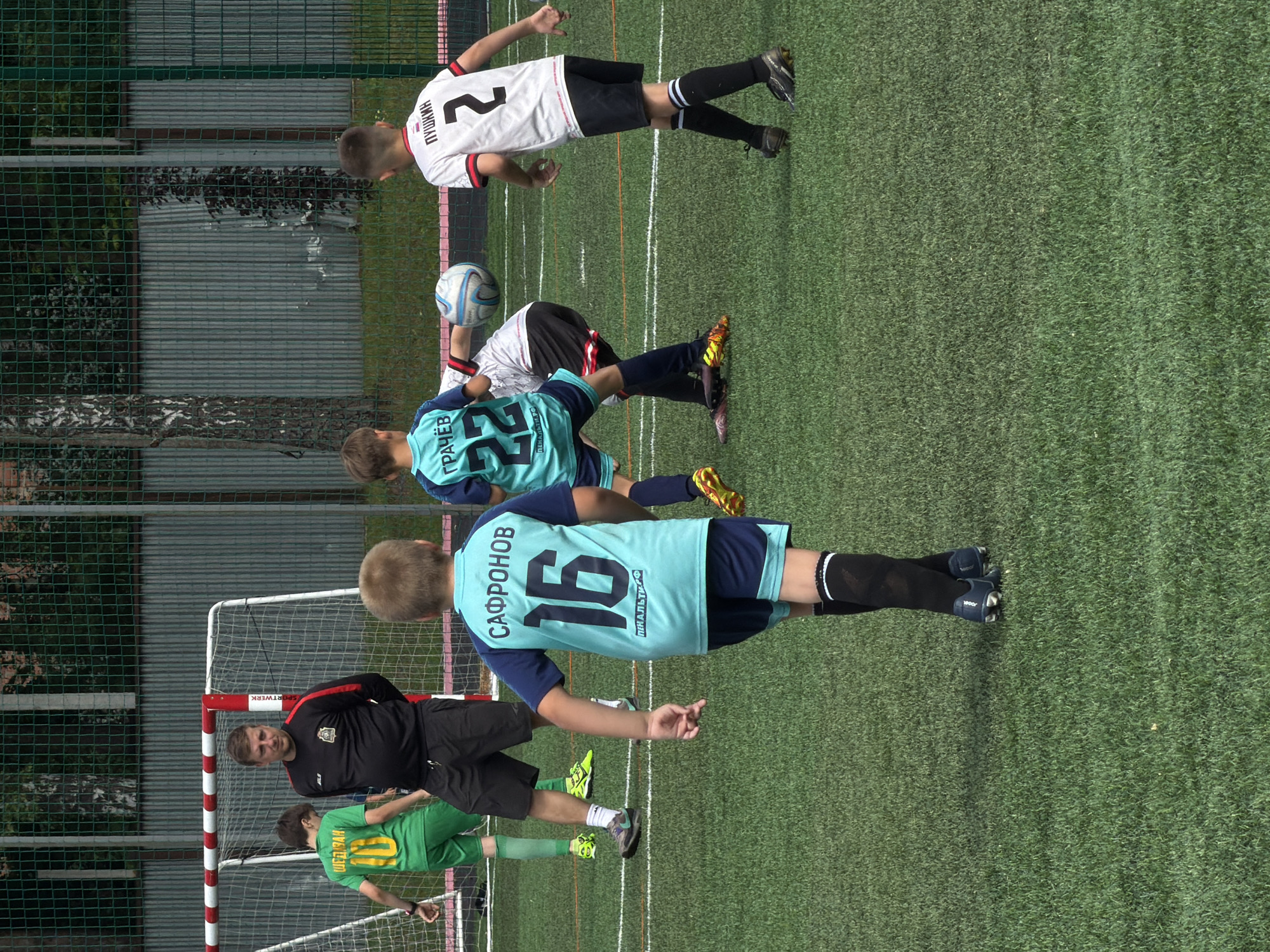 In Bykovo, Russia, a group of young boys and a coach are engaged in a soccer practice on an artificial turf field. The coach, wearing a black tracksuit with red accents, stands near a red and white goalpost. The players are in various soccer uniforms. One boy in a green jersey with the number "10" is visible on the left. Another boy in a teal jersey with "САФРОНОВ" and "16" on the back is in the foreground, facing away from the camera. To his right, a player in a white jersey with "ПУШКИН" and "2" is also visible. A boy in a teal jersey with "ГРАЧЁВ" and "22" is partially obscured, kicking a soccer ball. The background features a green metal fence, trees, and a grassy area. The overall setting suggests a typical youth sports event.