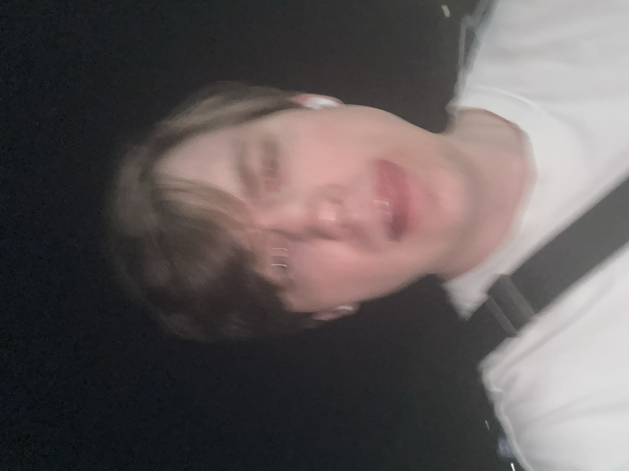 The image is a poorly lit, blurry selfie taken at night. A person with light-colored hair is in the foreground, their face partially obscured by the low light and focus. They are wearing a white t-shirt and white earbuds. A dark strap, possibly from a bag, is visible across their chest. The background is dark and indistinct, suggesting an outdoor or dimly lit indoor setting. There are no discernible landmarks or details that would indicate a specific location within the city of Dnipro, Ukraine.