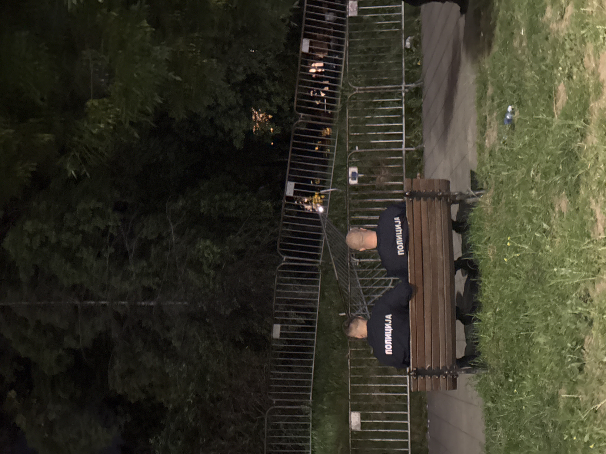 Two uniformed men labeled "ПОЛИЦИЈА" are sitting on a park bench in Belgrade, Serbia. Metal barriers surround the bench, possibly indicating a restricted area. The surrounding environment includes trees, grass, and a paved path. The scene appears to be set at night.