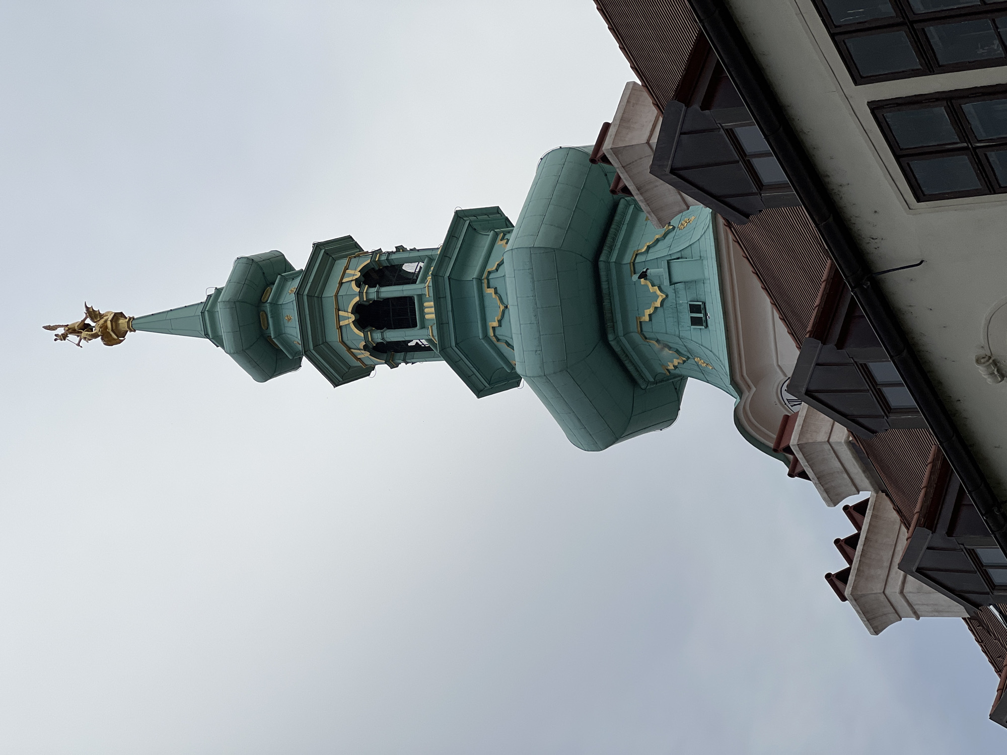 This is a low-angle, close-up photograph of a historical building's architectural features, specifically a tower and adjacent facade, under an overcast sky. The main subject is a distinctive green, multi-tiered tower, possibly a church steeple, with ornate gold accents. A small golden statue, appearing to be an angel or a winged figure, is visible at the very tip of the tower's spire.
To the right of the tower, a section of a building facade is visible, featuring dark-paned windows set within a light-colored wall. Several triangular roof-like structures with dark, slatted coverings jut out from this facade. The architectural style suggests a historical European setting. The cloudy sky indicates a daytime shot, likely with diffused light due to the weather.
The image's orientation is rotated, with the tower appearing to stretch upwards from the left side of the frame. The overall impression is one of historical grandeur and intricate craftsmanship. No people are visible, and no text is discernible in the image. The scene appears to be a quiet, possibly ancient, urban environment.