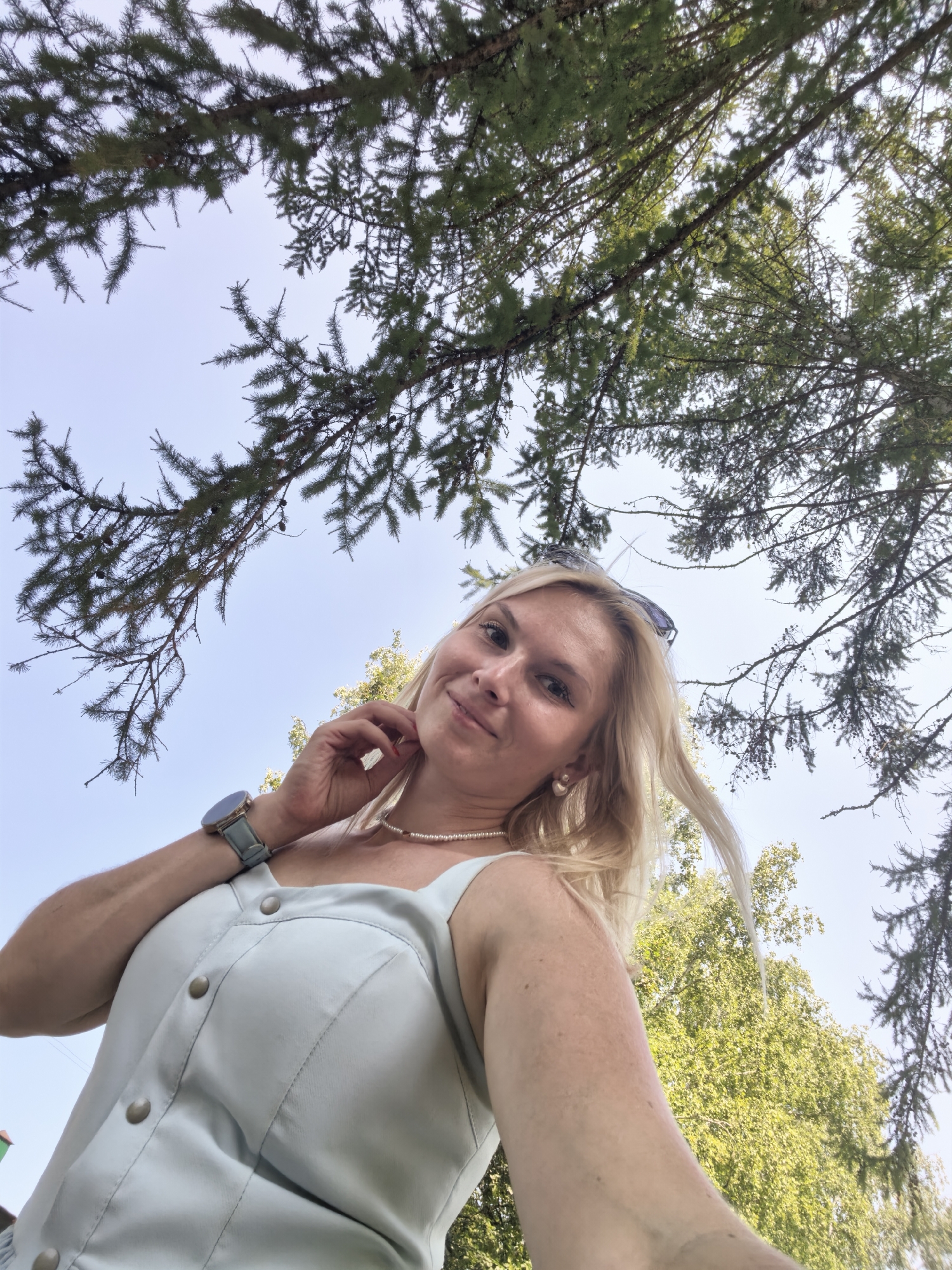 An outdoor, low-angle, eye-level, close-up, selfie-style photograph shows a blonde woman with fair skin and light-colored eyes looking at the camera. She has her right hand to her neck and is wearing a light blue tank top with three large buttons down the front, a pearl necklace, and a light blue watch on her left wrist. She is also wearing pearl earrings, and her hair is blonde and wavy, framing her face. In the background, there are green pine branches, a clear blue sky, and the tops of green trees. The photograph was taken outdoors in a location that could be Sterlitamak, Russia, as the presence of pine trees and deciduous trees in the background suggests a natural setting with a temperate climate.