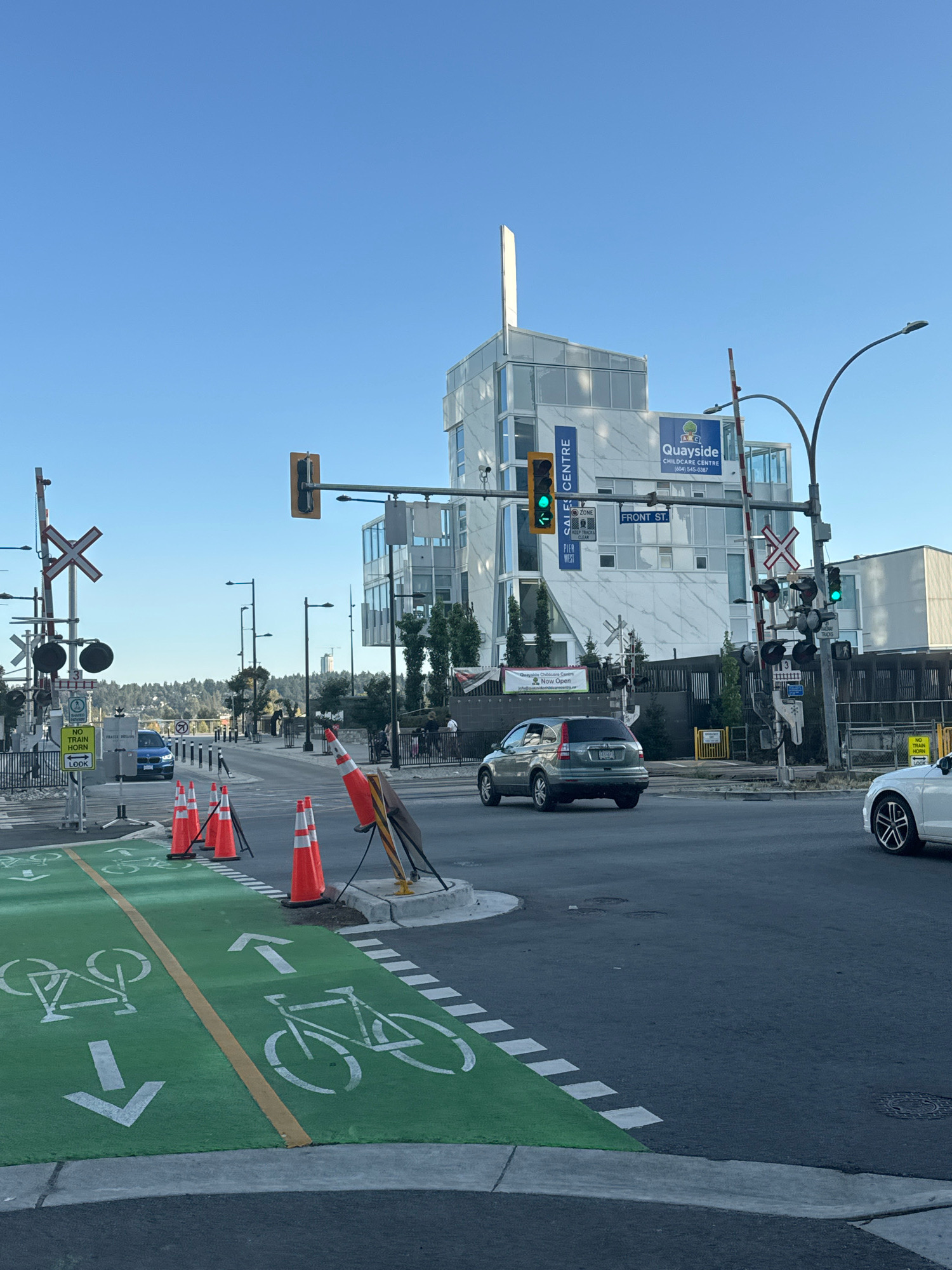 The image captures an urban street scene in New Westminster, Canada, featuring a prominent childcare center building and a railway crossing. The Quayside Childcare Centre, identifiable by its signage and modern architectural design with white marble-patterned cladding, is a central element. The building has large windows and a distinctive vertical white spire.
The intersection includes functional railway crossing signals with flashing red lights and barrier arms. Several traffic lights are visible, with one showing a green light for vehicular traffic. The road surface is asphalt, with newly painted green bike lanes clearly demarcated by white dashed lines and bicycle symbols. Arrows indicate traffic flow for cyclists, with one lane showing an arrow pointing forward and another showing an arrow pointing backward.
Orange traffic cones are positioned near the bike lane, suggesting temporary traffic management or construction. A silver SUV is turning at the intersection, and a white car is visible to the right. A blue vehicle is also present further down the road. In the background, a cityscape with buildings and a distant treeline is visible under a clear blue sky. The overall environment suggests an active urban area with infrastructure for both vehicles and cyclists, as well as a pedestrian-friendly approach due to the bike lanes.