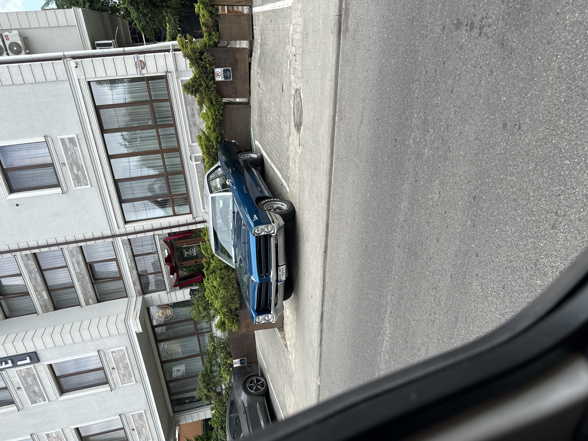 The image displays a street view in Durlești, Moldova, featuring a blue vintage Pontiac parked on a cobblestone lot in front of a modern building. The building, with its light-colored facade and large windows, appears to be a commercial establishment, possibly a hotel or restaurant, as suggested by the "HOTEL" sign visible on the upper corner and a "COFFEE MORE" sign near the entrance. To the left, a dark gray modern sports car is parked in an adjacent spot. The foreground is dominated by the asphalt road, partially obscured by a dark, out-of-focus element, likely the interior of a vehicle from which the photo was taken. Lush green bushes are planted in rectangular planters flanking the parking area, providing a natural contrast to the urban architecture. A parking sign is affixed to one of the planters. The sky is overcast.
