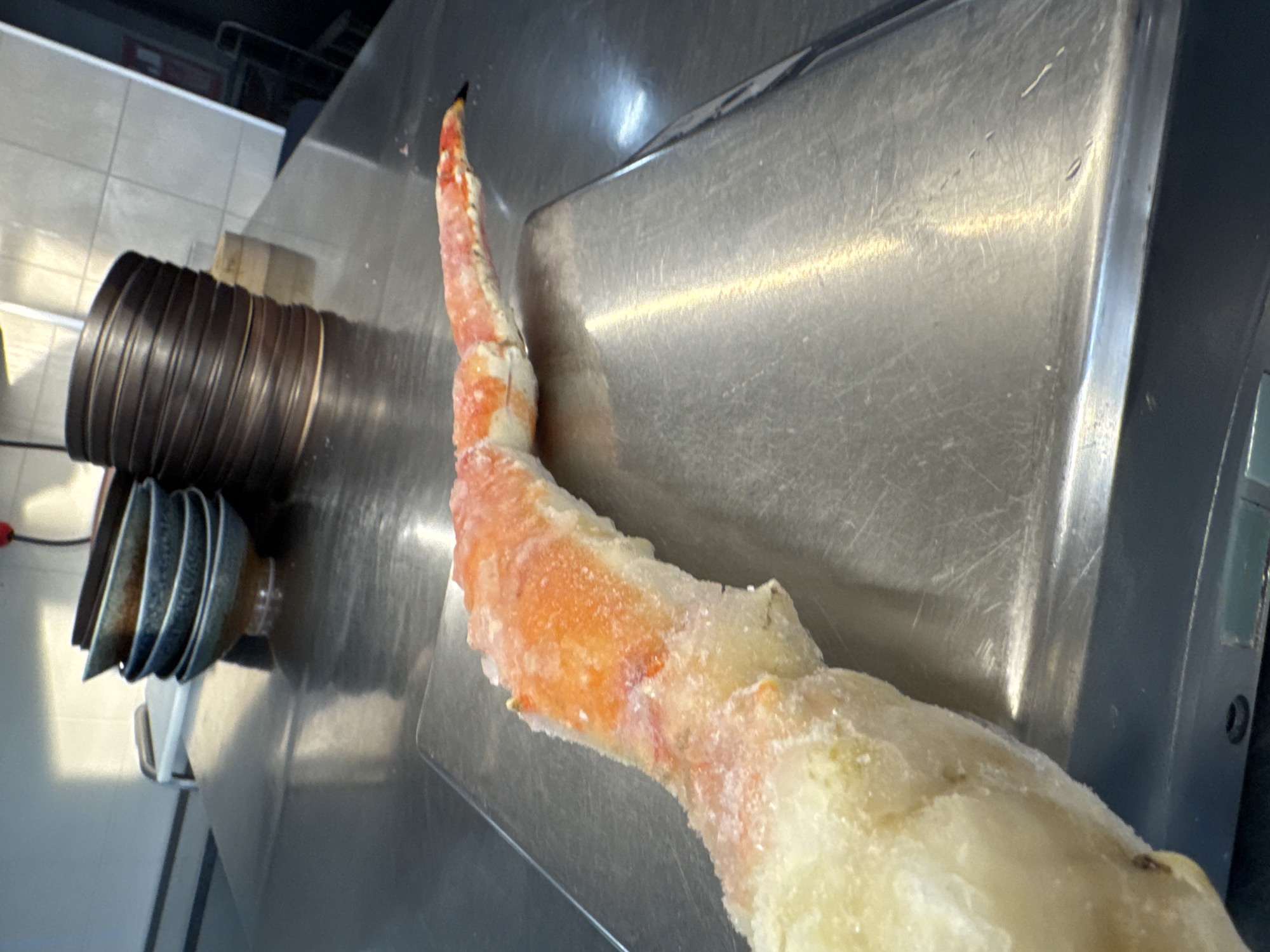 The image shows a frozen crab leg resting on a stainless steel surface. To the left of the crab leg, a stack of bowls is visible. The background includes a wall with light-colored tiles. The image was likely taken indoors, possibly in a kitchen or food preparation area. There is no immediate evidence to confirm the location is in Sofia, Bulgaria.