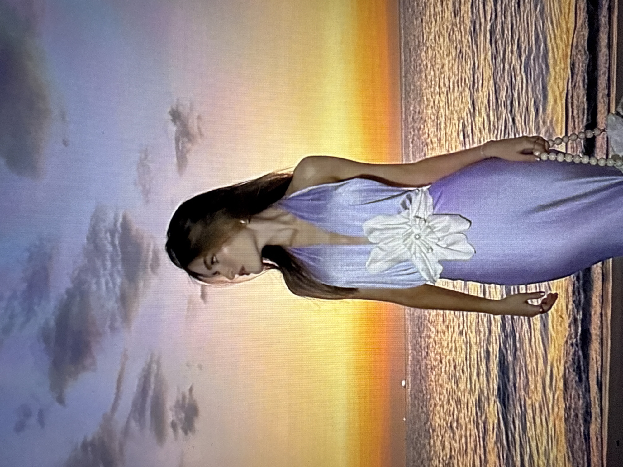 The image shows a young woman standing on a beach in front of a sunset. She is wearing a light purple, halter-neck dress with a large white flower embellishment on the bodice. The dress has a plunging neckline. The woman is looking to her right, away from the camera. Her hair is long and brown. In her right hand, she is holding a beaded bag. The ocean is behind her, with gentle waves lapping at the shore. The sky is filled with colorful clouds in shades of orange, yellow, and purple, indicating a sunset or sunrise. There are a few distant lights visible on the horizon over the water. The overall scene is serene and picturesque. The image is not a photograph of Găvănoasa, Moldova, as it depicts a beach and sunset, which are not characteristic of that specific city.