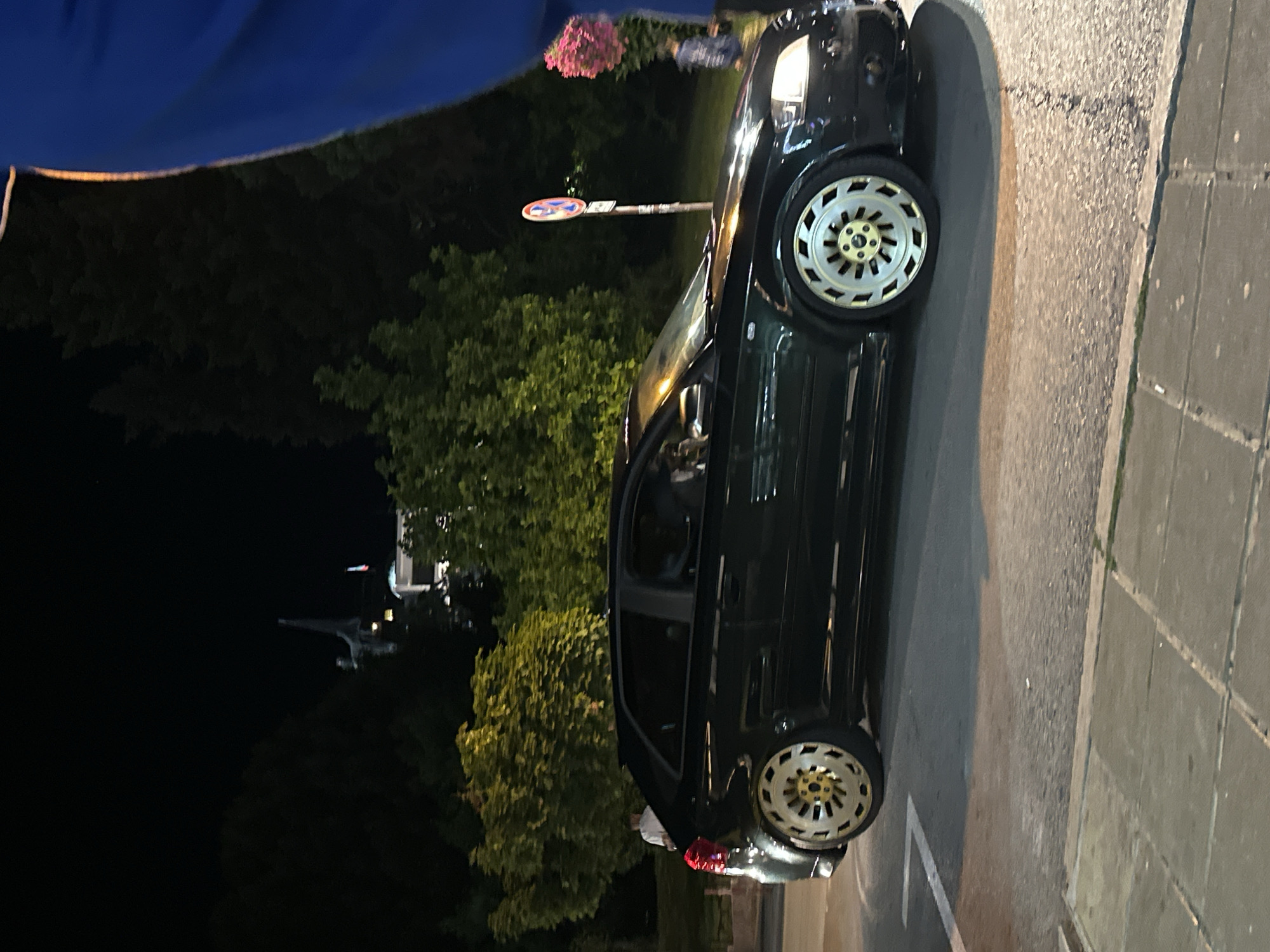 A black Audi A3 is parked on a street at night. The car has custom gold rims. In the background, there are trees and buildings, including a building with a statue on top and a Bulgarian flag. A person in a blue shirt is partially visible on the right side of the frame. The street is paved with asphalt and concrete tiles. There is a traffic sign in the background. The lighting suggests it is evening or night.