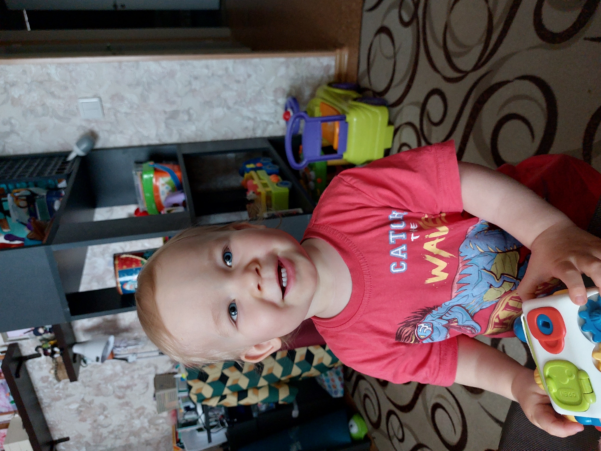 A young child with blonde hair and blue eyes is playing with a toy. The child is wearing a red t-shirt with a dinosaur graphic. The child is sitting on a brown rug with a swirl pattern. Behind the child is a dark gray shelving unit with various toys and objects on the shelves. A light switch is visible on the patterned wall. The location is Chaikovsky, Russia.