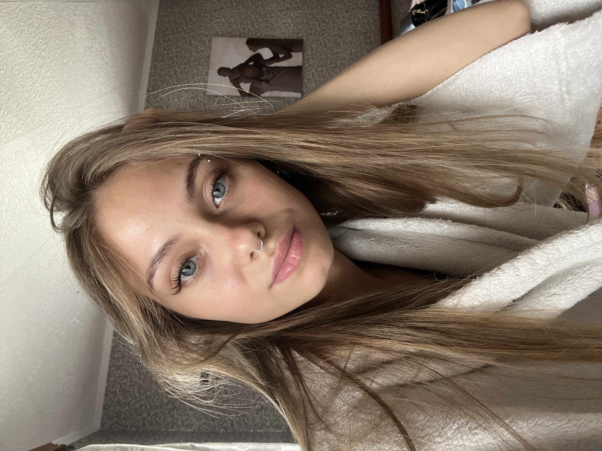 A young adult female with long, straight light brown hair is depicted in a close-up, waist-up view within an indoor setting. Her head is tilted slightly to the left, and she gazes directly into the camera with bright blue eyes, enhanced by prominent artificial eyelashes on her upper lids. Her complexion appears clear. She has a horizontal barbell piercing located above her left eyebrow (viewer's right) and a decorative L-shaped nose piercing with small white stones on her left nostril. Her lips are full and naturally pink, slightly parted. She is wrapped in a light-colored, possibly off-white or cream, textured, fluffy garment resembling a bathrobe or blanket. Her right arm (viewer's left) is bent, with her hand or forearm resting near her head, partially obscured by her hair.
The background consists of a light gray or beige patterned wallpaper on the wall behind her. A white, textured ceiling is visible at the top of the frame. On the right side of the image, a framed picture or poster is mounted on the wall, depicting three figures: one adult carrying a baby on their shoulders, and another adult standing to the right. The figures are somewhat indistinct or silhouetted. On the far left, a glimpse of a lighter, possibly striped, wall or curtain is visible. The lighting suggests an indoor environment, possibly illuminated by natural light from an unseen window. The specific location, including a city like Фоки, Russia, cannot be determined from the visual information provided within the image.