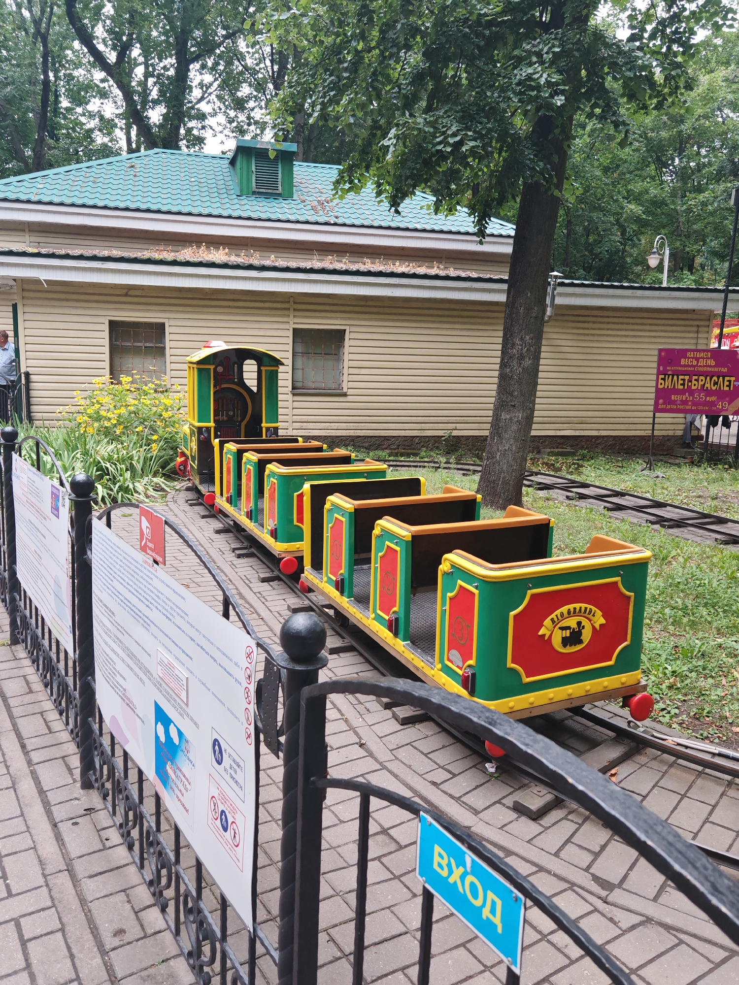 The image depicts a miniature train parked on tracks in an outdoor setting. The train is colorful, with green and red carriages adorned with yellow trim. The leading car, resembling a locomotive, also features green and yellow colors with red accents. The train appears to be part of an amusement park or recreational area, indicated by the presence of a fence, informational signs, and a building in the background. The building has a light-colored facade and a green, wavy roof, with windows visible. Trees with lush green foliage surround the area, suggesting a park environment. The ground is paved with interlocking stones, and there is grass and other vegetation visible. On the fence, there are several signs with text and graphics. One sign, in blue, has the word "ВХОД" (ENTRANCE) written in yellow Cyrillic letters. Another sign, pink in color, displays information about a "BILET-BRASLET" (TICKET-BRACELET). A person is partially visible on the left side of the image, standing near the building. The overall scene suggests a bright, possibly overcast day, with ample greenery and a playful, family-friendly atmosphere. The presence of Cyrillic script on the signs strongly indicates the location is in a country that uses this alphabet, such as Belarus, Russia, or Ukraine. Given the prompt specifies the city of Homyel, Belarus, it is reasonable to infer this is the location.