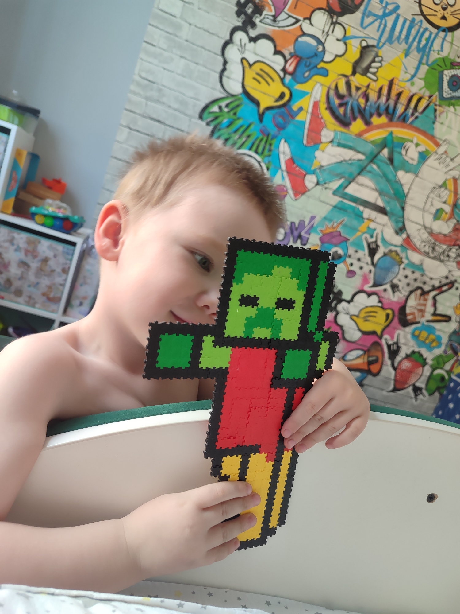 A young, light-skinned, blond-haired child is holding a pixel art figure of a character resembling a Minecraft zombie. The figure is green on the head and arms, red on the torso, and yellow on the legs, all outlined in black. The child is holding the figure in front of them, looking down and to the side. The child is in an indoor setting, likely a child's bedroom. Behind the child is a wall covered in colorful graffiti-style art. To the left, slightly out of focus, are shelves containing toys and other items. The location is Izhevsk, Russia.