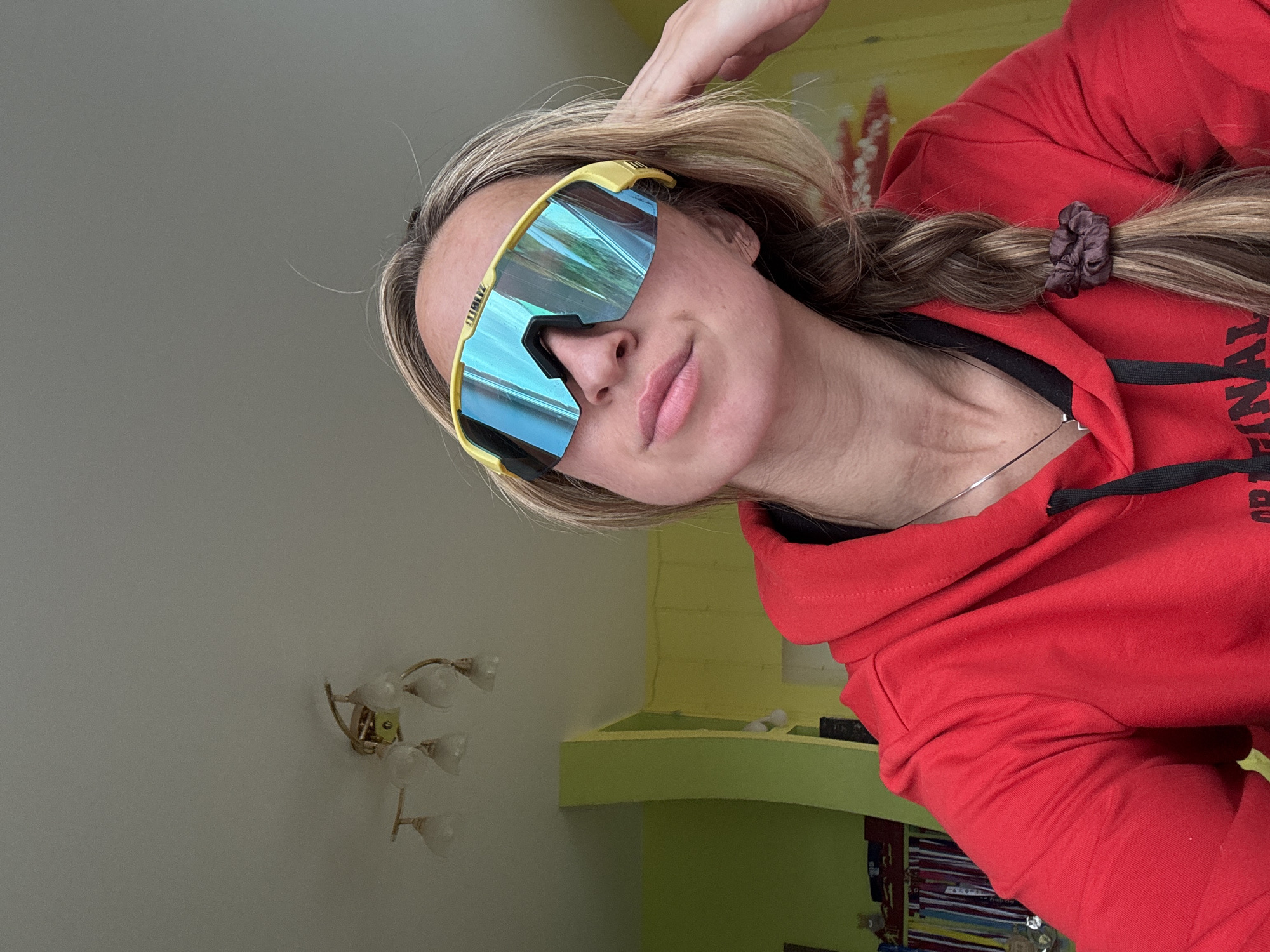 A young woman with blonde hair in a braid is posing for a selfie. She is wearing a bright red hooded sweatshirt and large, futuristic yellow and blue sunglasses. The background appears to be an indoor setting, with a wall painted a bright yellow and a green shelf. There are some decorative wall lights visible on the left side of the image. The woman's hand is resting on her head, and she has a slight smile on her face. The overall impression is casual and playful. Text is visible on her sweatshirt but is cut off and illegible. The location is given as Prikamskiy, Russia, but there are no specific visual cues to confirm this. The lighting suggests it is daytime.