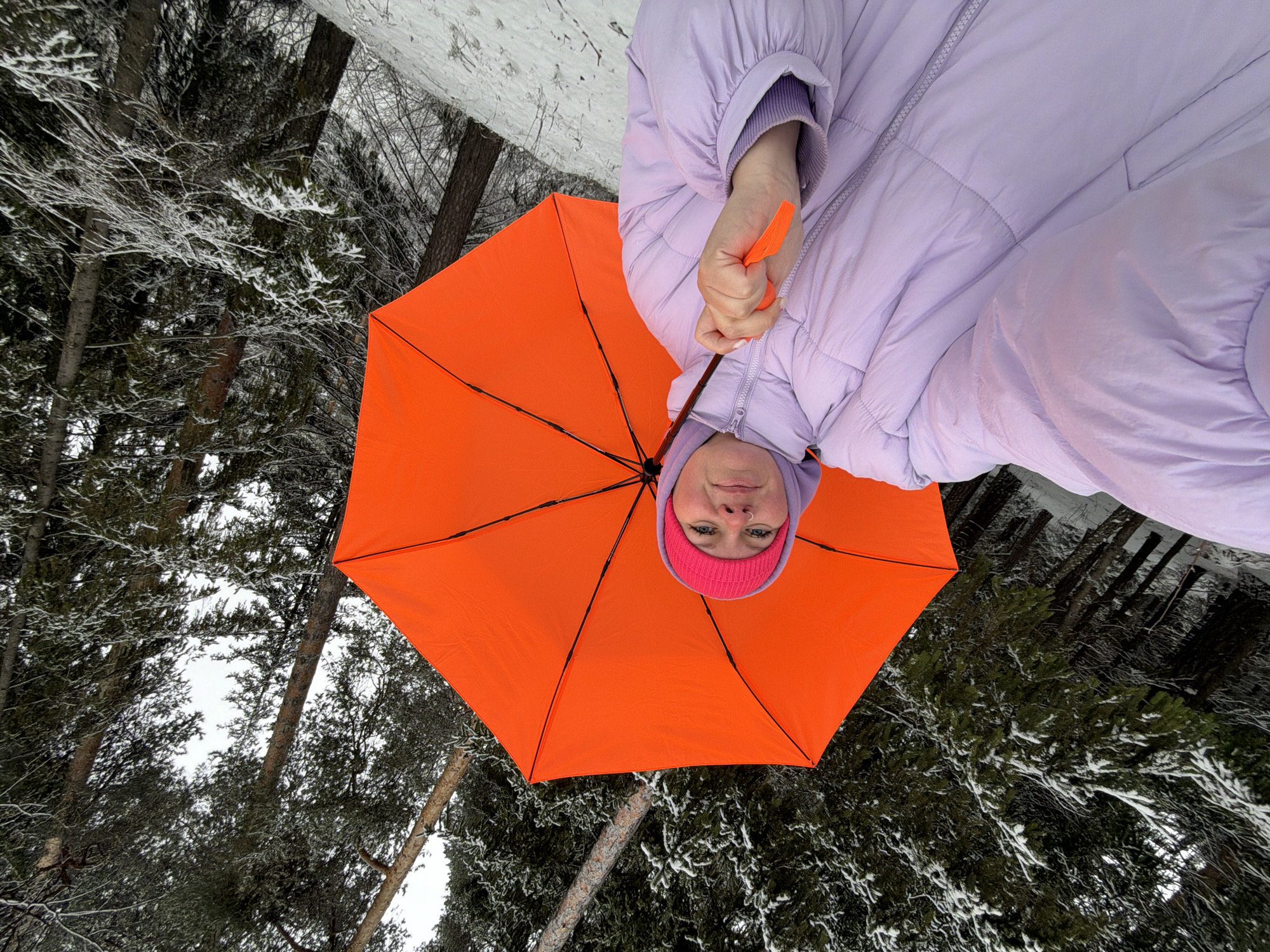 A person is depicted in a low-angle, upside-down perspective in a winter scene. The individual wears a lavender puffer jacket and a pink beanie. They are holding an open, bright orange umbrella. The setting appears to be a snow-covered forest with trees visible in the background, some dusted with snow. The image was taken in the city of Chaikovsky, Russia.
