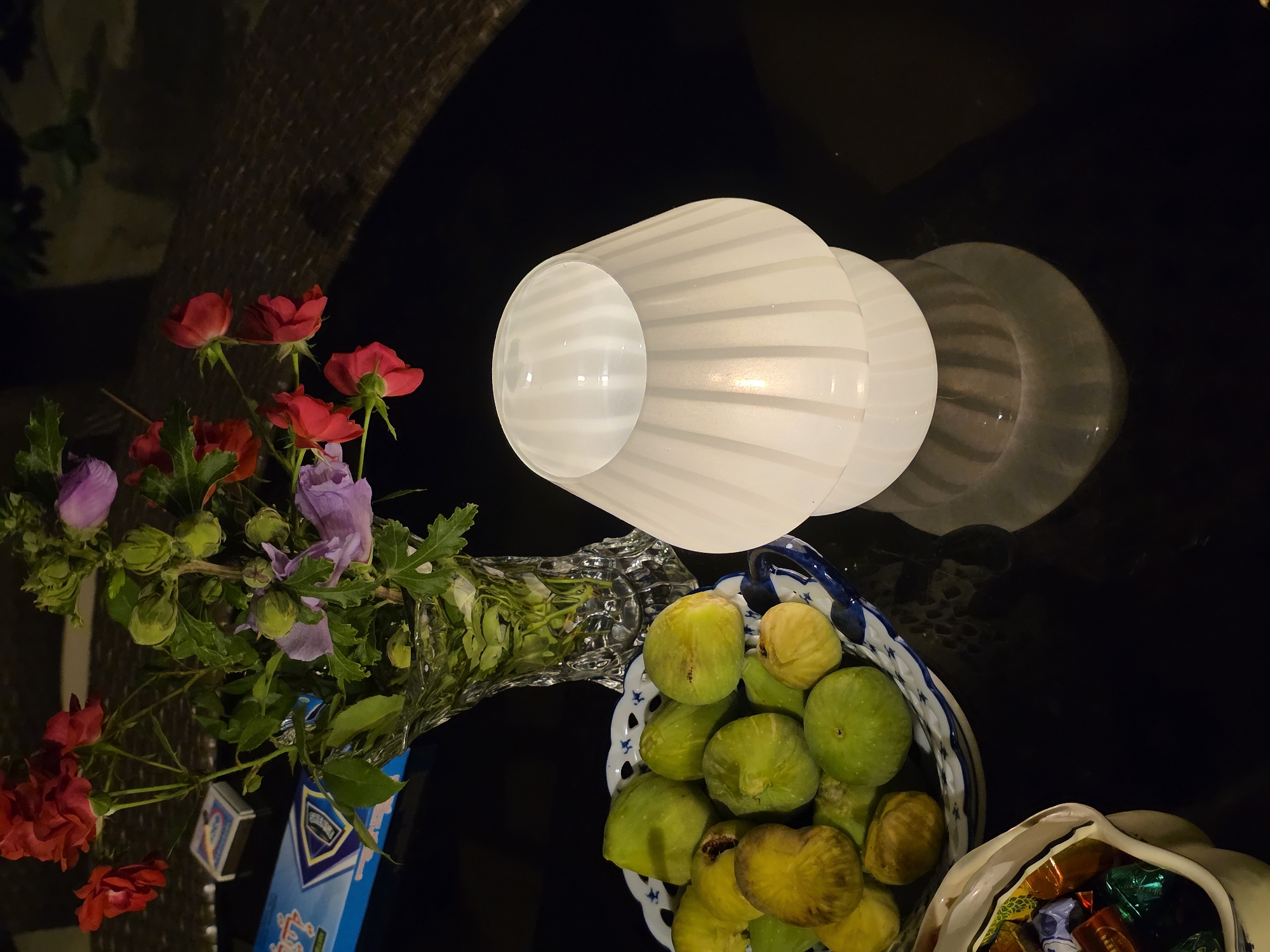 The image depicts a close-up, high-angle shot of a dark, glossy table at night. On the table, a clear glass vase holds a bouquet of red roses and purple hibiscus flowers, with some unopened buds. To the left of the vase, a blue box with white and gold markings sits next to a small box of matches. Below the vase, a blue and white patterned bowl overflows with green and yellowish-brown figs. To the right of the figs, a white mushroom-shaped lamp with vertical ridges emits a warm glow from within, casting a reflection on the table surface. In the bottom left corner, a white rectangular dish holds an assortment of individually wrapped candies. The background appears to be an outdoor setting, possibly a patio or garden, with the faint texture of woven furniture visible in the upper right corner. The scene is likely located in Buzovna, Azerbaijan, given the context of the request.