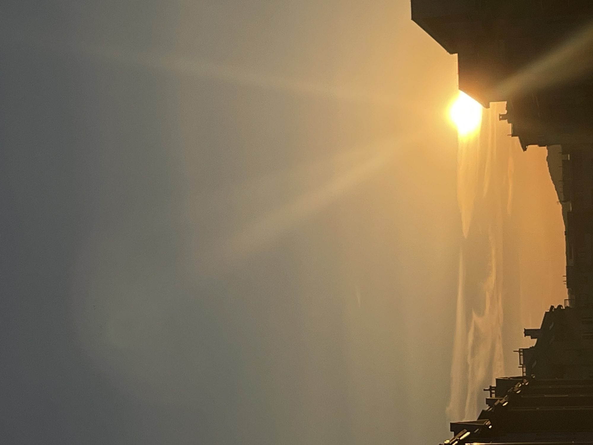 The image captures a sunset over a cityscape. Silhouetted buildings, some with chimneys, line the bottom of the frame. The sun is a bright, glowing orb positioned in the right half of the image, partially obscured by a building. Rays of sunlight fan out from the sun, creating a dramatic effect against the soft orange and gray hues of the sky. Wispy clouds are visible in the middle ground. The scene appears to be an urban environment.