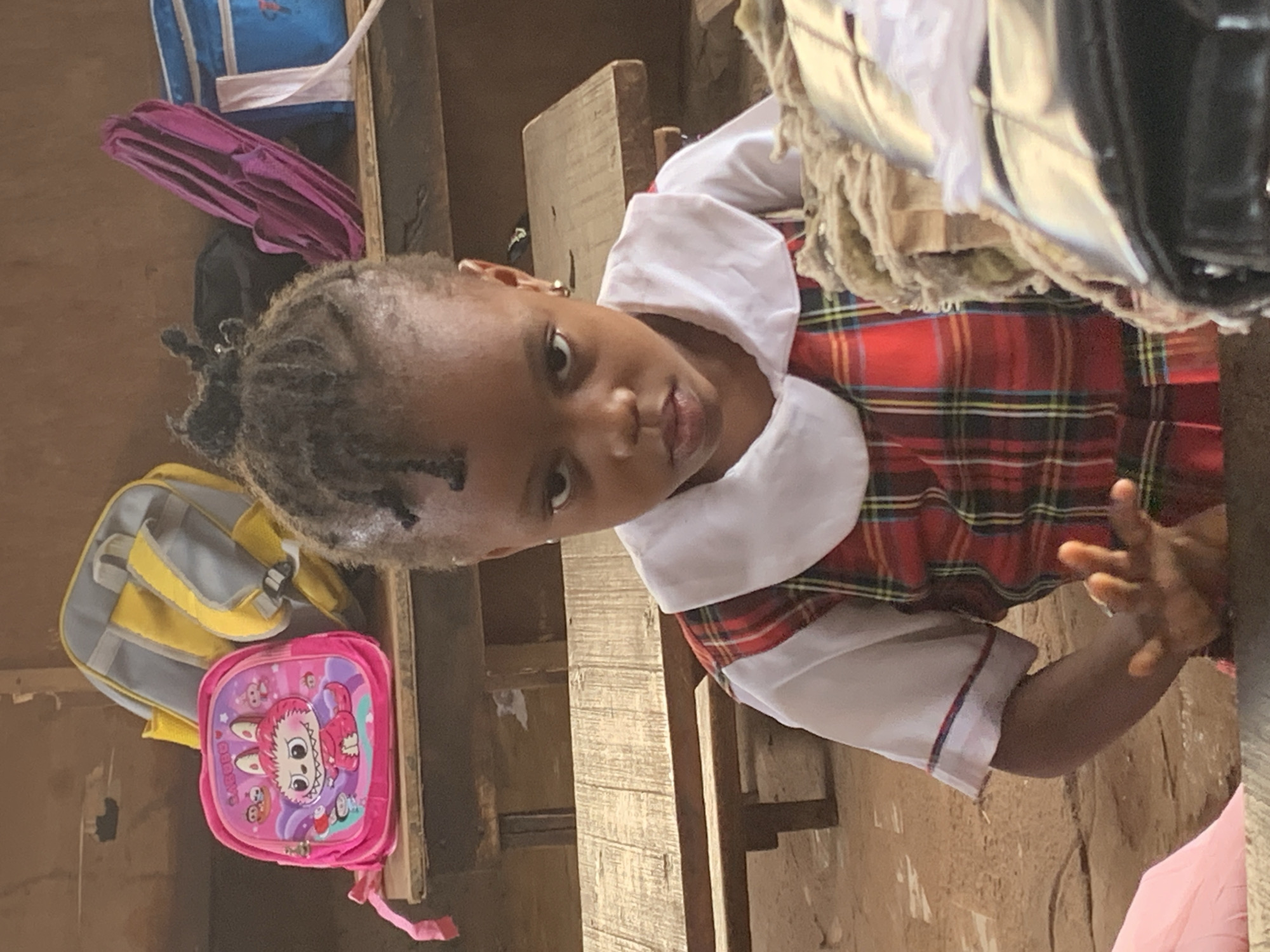 A young girl, presumably in a school uniform with a white collar and red plaid pattern, is the primary focus of the image. She is looking directly at the camera. Behind her, there are school backpacks, one pink with a cartoon character and another yellow and gray. There appears to be a wooden structure, potentially a wall or shelving unit, in the background. The location is likely Ukwu-Abwa, Nigeria.