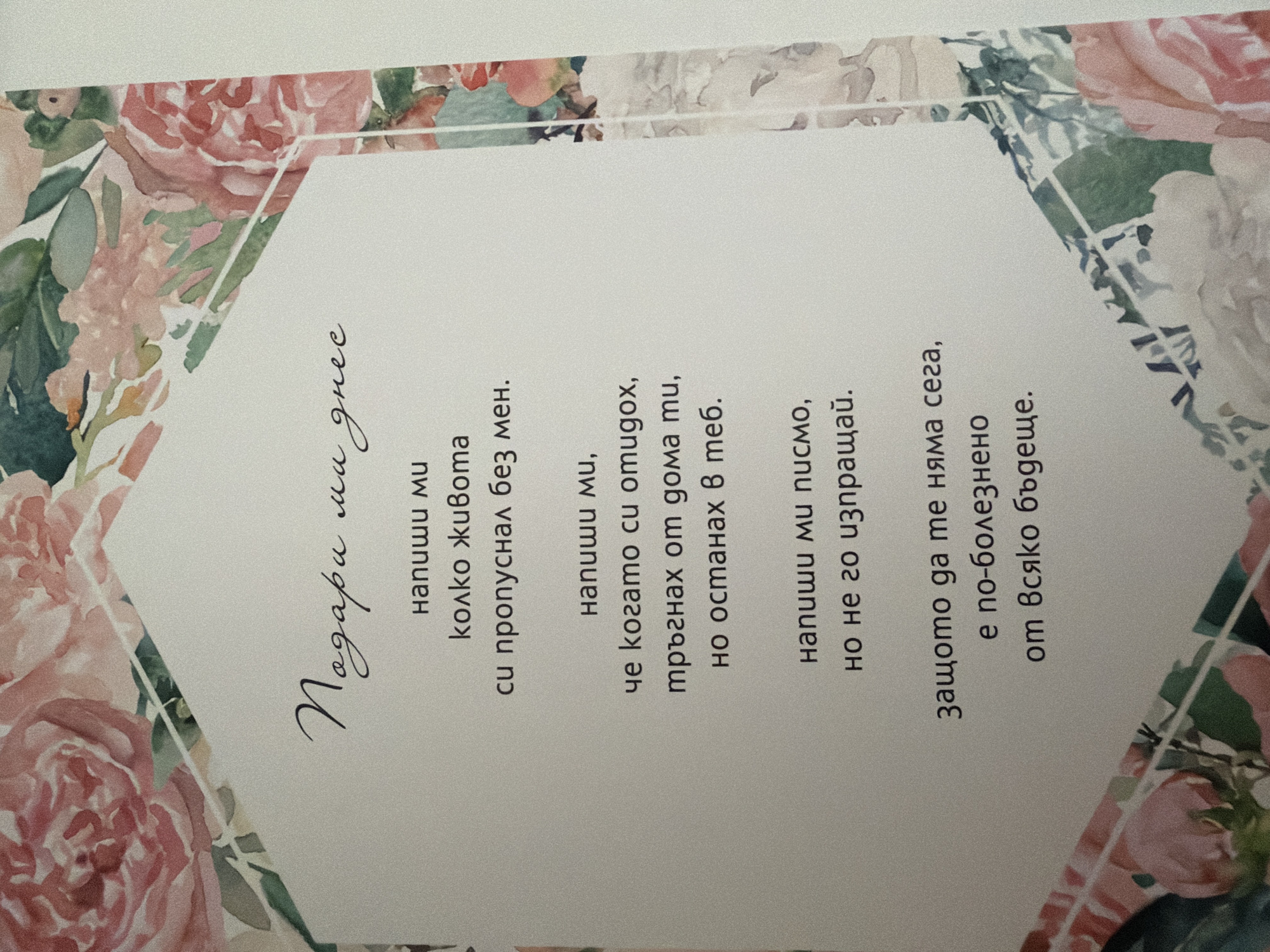 The image displays a greeting card with floral motifs, featuring text in Bulgarian. The text reads:
"Подари ми днес
напиши ми
колко живота
си пропуснал без мен.
напиши ми,
че когато си отидох,
тръгнах от дома ти,
но останах в теб.
напиши ми писмо,
но не го изпращай.
защото да те няма сега,
е по-болезнено
от всяко бъдеще."
There are no people, actions, or interactions visible in the image. The environment is that of a flat surface, likely a table or desk, where the card is placed. The image does not contain any information or visual cues that would allow for its location to be determined within Pazardzhik, Bulgaria, or any other city.