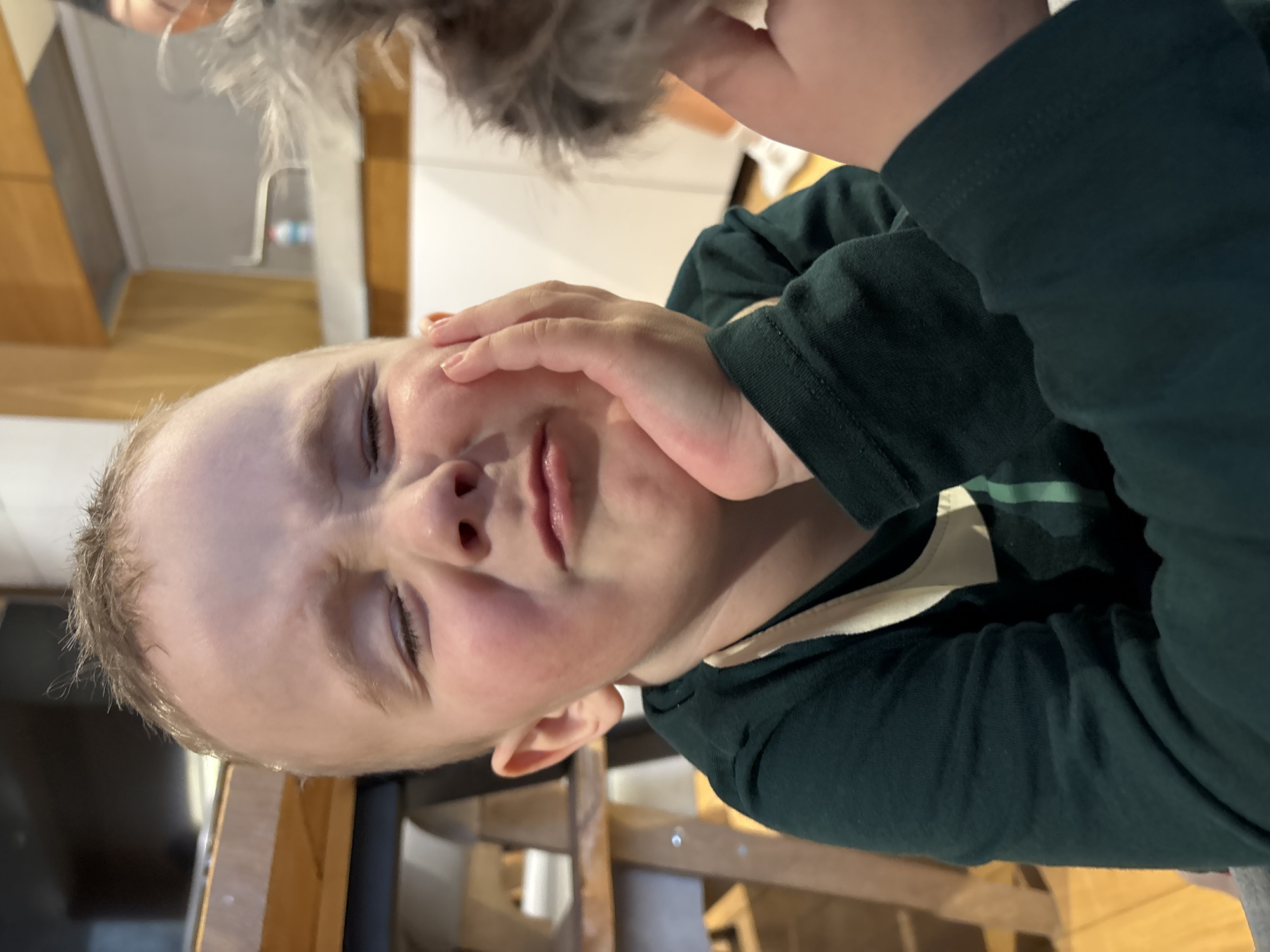 An indoor scene in Chaikovsky, Russia, depicts a young child with fair skin and light brown hair captured in a close-up, chest-up shot. The child's eyes are tightly closed or squinted, eyebrows are furrowed, and the mouth is pursed downward, indicating a grimacing or distressed expression. The child's left hand is pressed against their left cheek. They are wearing a dark green long-sleeved top with a lighter-colored trim visible at the neckline. The child's right hand, partially visible, holds a blurry, greyish-brown, textured object, possibly a stuffed animal. The background to the left features wooden furniture or counter surfaces. The background to the right reveals a kitchen area with white and light wood cabinets, a metallic range hood, a faucet, and a small blue and white container on a countertop.