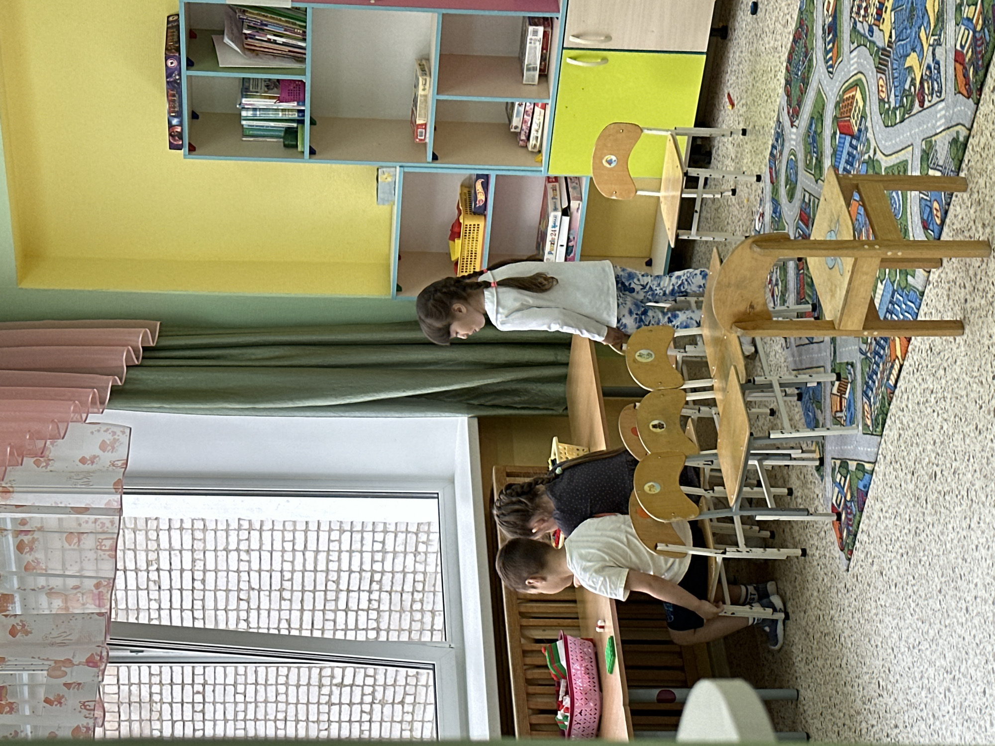 The image depicts an indoor scene, likely a classroom or playroom, with a focus on young children and educational materials. The room is brightly lit and decorated with colorful elements.
In the foreground, several children are visible. A young boy, wearing a white t-shirt and dark shorts, is seated at a wooden table, facing away from the camera. To his right, a young girl with braided hair, dressed in a dark top, is also seated at the table. Further to the right, another young girl in a white long-sleeved shirt and patterned pants stands, looking towards the left. In front of the seated children are several small wooden chairs and a rug with a city or road pattern.
The table they are seated at appears to be a low-height table designed for children. On the table, there are baskets containing what appear to be toys or learning materials, and some scattered small objects.
The background features a large window with a brick wall visible outside. Curtains, one pink and one green, frame the window. To the right, a colorful, multi-shelf unit is filled with books, toys, and boxes, suggesting it is a storage and display area for educational resources. The walls of the room are painted in bright yellow and light green.
The overall impression is that of a learning environment for young children, possibly a kindergarten or preschool. The presence of children, toys, books, and child-sized furniture points towards this interpretation.
There is no information available in the image to ascertain the specific location within the city of Chaikovsky, Russia.