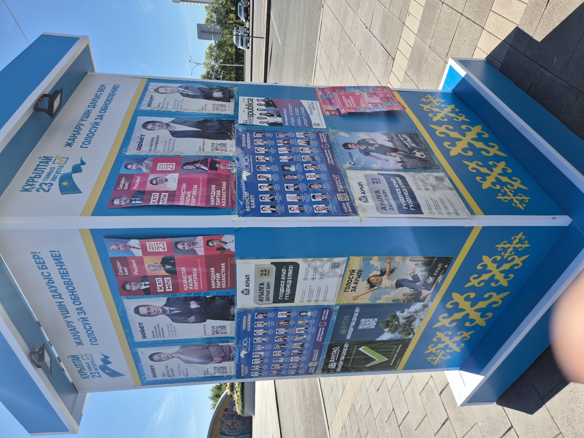 This image shows a bright blue and yellow kiosk adorned with political posters in Astana, Kazakhstan. The kiosk has a white top with an integrated light fixture. The posters are primarily in the Kazakh language, with some Russian text, and feature portraits of various individuals, likely candidates in an election. Several posters mention "23 тамыз" (August 23rd) and the slogan "Жаңару үшін дауыс бер!" (Vote for renewal!). One poster displays the text "ҚҰРЫЛТАЙ" (Kurultai), which can refer to a congress or assembly. There are also party logos and slogans. The kiosk is situated outdoors on a paved area, with trees and a building visible in the background. The sky is clear and blue, indicating a sunny day. The kiosk itself is decorated with traditional Kazakh patterns in yellow.