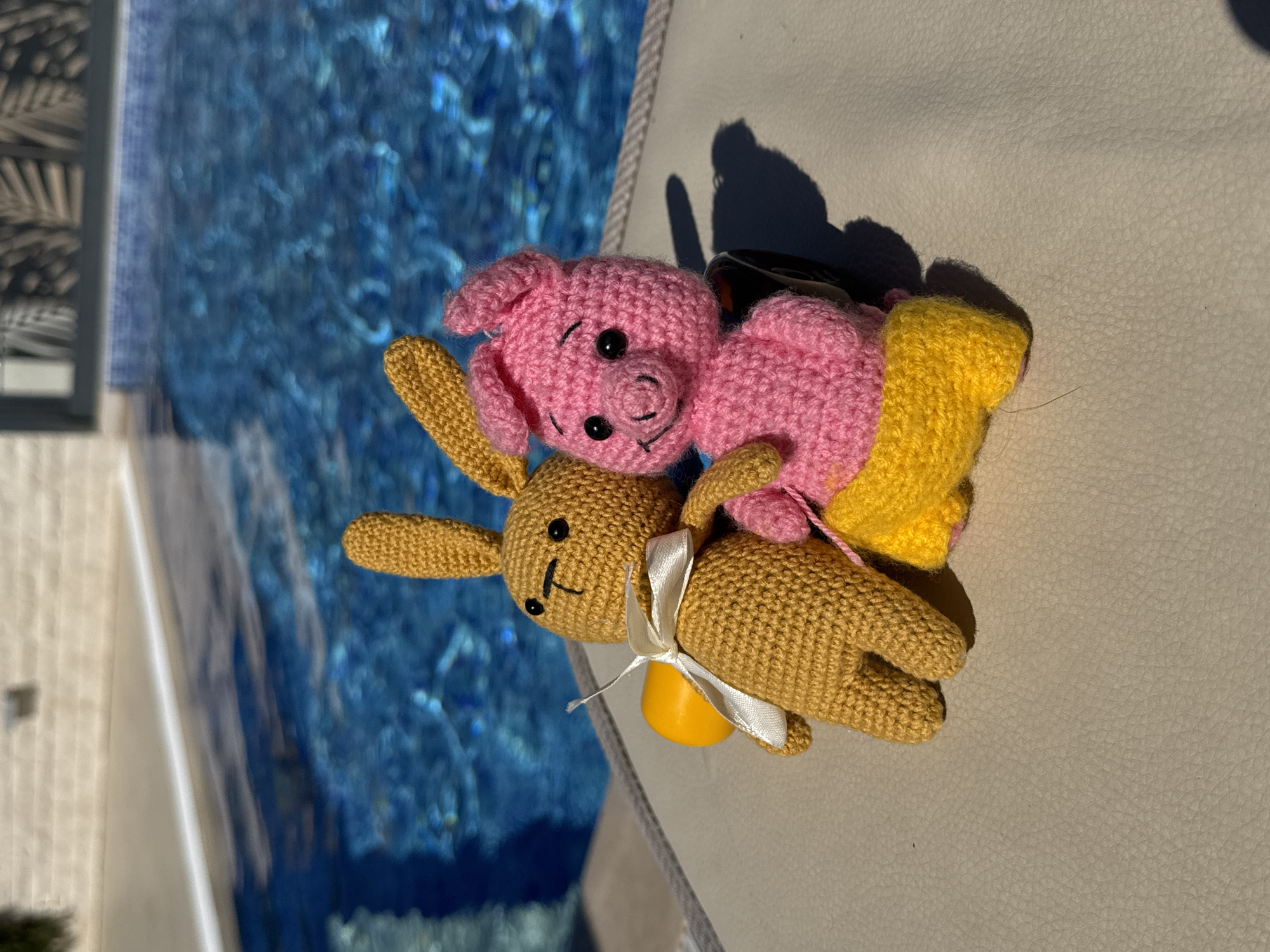 A top-down shot shows two crocheted toys—a pig and a rabbit—lying next to a swimming pool. The pig is pink with black eyes and a curled snout. It is partially covered by a yellow crocheted item and appears to be resting on top of the rabbit. The rabbit is a tan color and has a white ribbon tied around its neck, secured to a yellow item. The pool is blue with blurred reflections visible. The toys and pool are located on a surface that appears to be white or light grey. The scene is brightly lit with a shadow cast to the right of the toys, indicating a daytime setting. The location is Đenovići, Montenegro, inferred from the presence of a swimming pool and materials suggesting a pool-side environment.