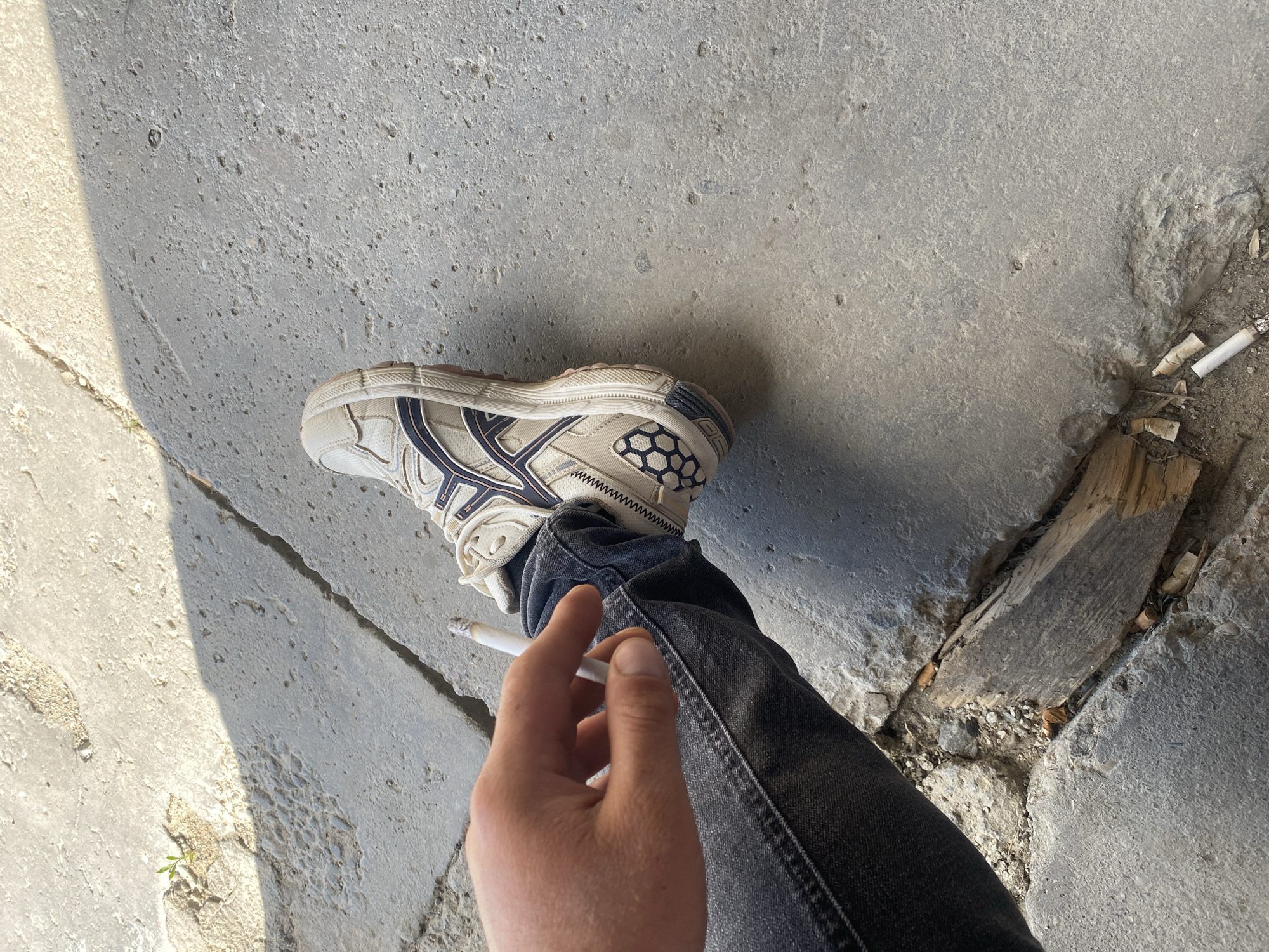 The image features a person's lower body, specifically their right leg clad in dark denim jeans and a white athletic shoe with navy blue accents, placed on a cracked concrete surface. A lit cigarette is held between the person's thumb and index finger, near the shoe. In the background, several discarded cigarette butts are visible on the ground, along with a piece of weathered wood. The lighting suggests it is daytime, with a shadow cast across the concrete, indicating sunlight. The scene appears to be outdoors, possibly an urban or semi-urban environment, given the concrete and scattered debris. No text is visible in the image. The context provided places the location as Balta, Ukraine.