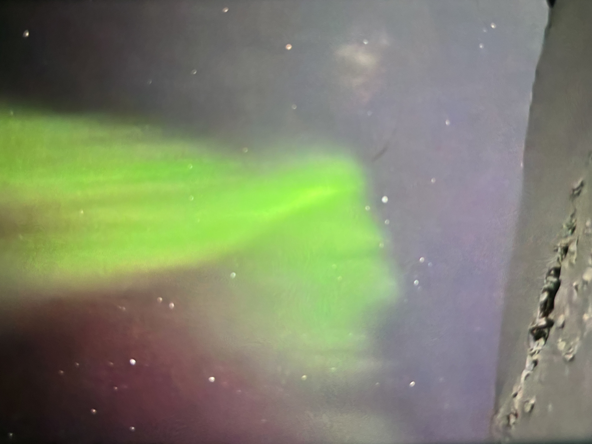 The image depicts a landscape scene under a night sky illuminated by a vibrant green aurora borealis. The aurora displays streaks of light in shades of green and subtle hints of purple. Scattered throughout the sky are numerous small, bright points, likely stars. In the foreground, a dark, uneven terrain with some lighter patches suggests a natural landscape, possibly mountainous or hilly, with some snow or ice. The overall impression is a serene and natural environment. The location is identified as Unknown, Unknown.