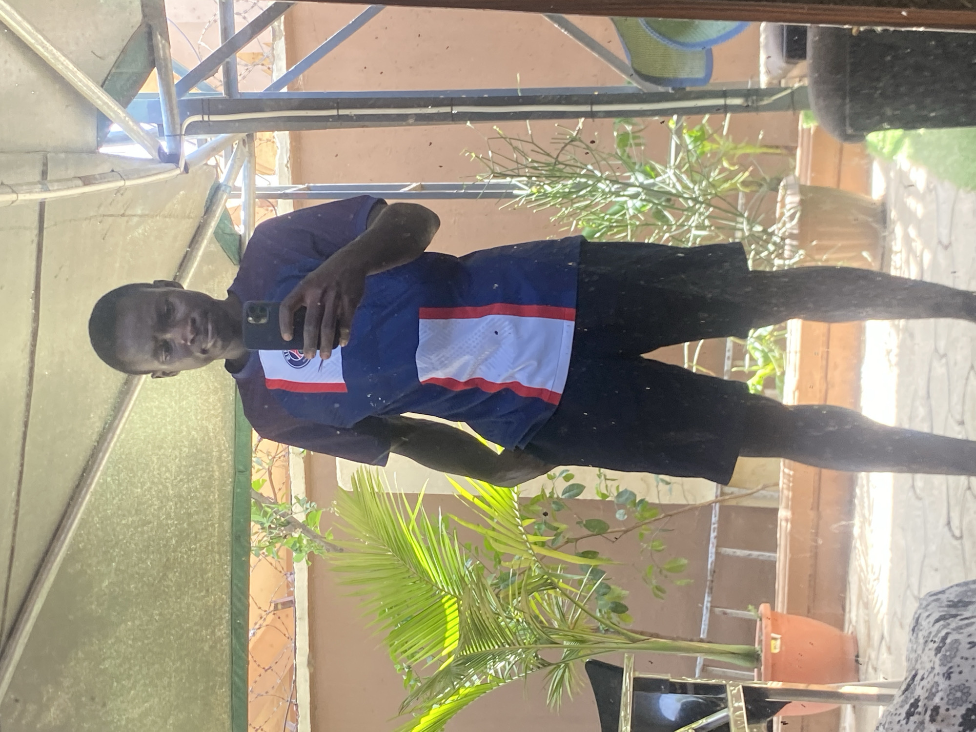 A dark-skinned man is taking a selfie in front of a reflective surface. The man is wearing a blue and white shirt and dark shorts. The background contains plants, a building with a peach-colored exterior, and a metal structure. Based on the location in Barden Yana, Nigeria, the scene is likely a residential or outdoor area.
