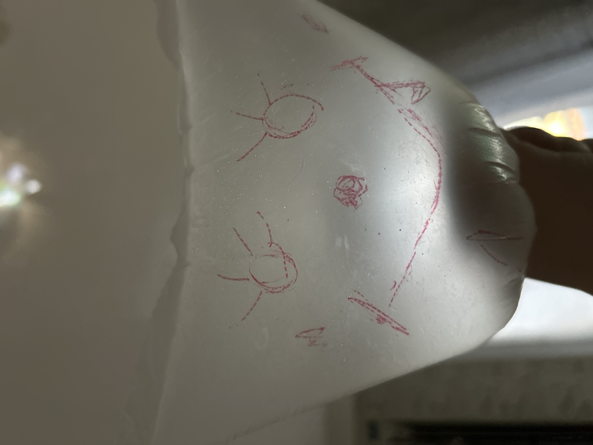 The image shows a white, translucent material, possibly plastic sheeting or a balloon, with red crayon or marker drawings on it. The material is held in a hand, which is partially visible on the right side of the frame. The drawings are rudimentary and appear to be abstract shapes and figures. One drawing on the left resembles a stylized sun with an oval center and radiating lines. Another drawing on the right is more complex, with lines that suggest a figure or vehicle with angular shapes. There are also smaller scribbles and dashed lines scattered across the surface.
The lighting in the image is diffused, with some bright highlights suggesting a light source from behind or above the material. The background is out of focus and appears to be an indoor setting with some light coming through a window or opening, casting shadows. There is no visible text in the image. The overall impression is one of a simple, possibly childlike artistic endeavor on an unusual medium. The translucent nature of the material allows some light to pass through, creating a soft glow and highlighting the texture of the surface, which appears slightly creased and possibly bumpy. The focus is primarily on the drawings and the material they are on.