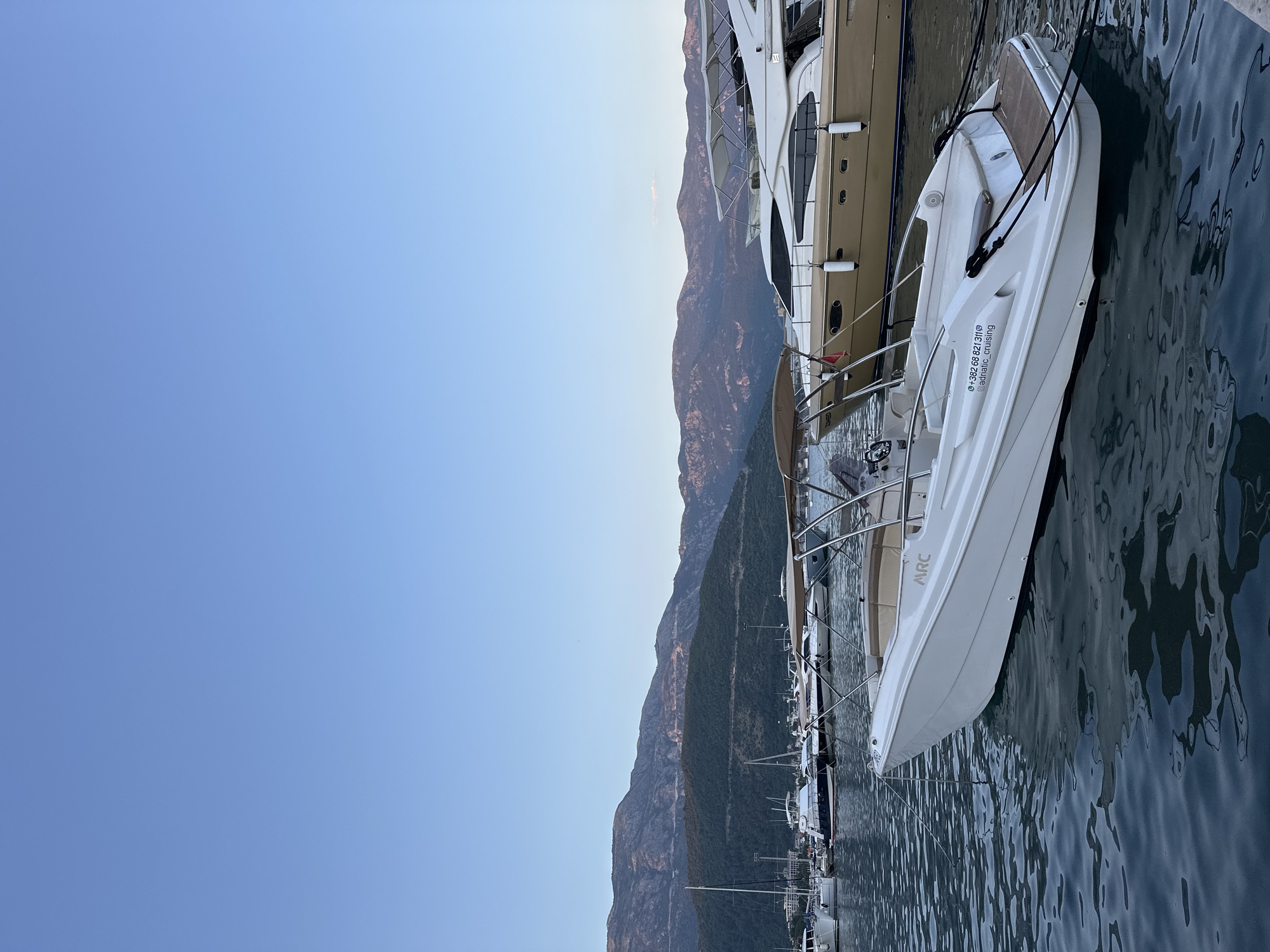 In Budva, Montenegro, a white motorboat with a tan canopy is moored in a harbor, its bow pointing towards the viewer. The boat has "ARC" logo on its side and contact information, "+382 68 821 311 Adriatic_cruising", on its stern. It is tied with black ropes to the dock. Behind this boat, a larger, tan yacht with white accents is also moored. The background is dominated by a clear blue sky and a range of mountainous terrain, with some greenery visible on the slopes. Several other boats, including sailboats, are docked further back in the harbor. The water is calm with gentle ripples. The scene appears to be at dusk or early morning due to the soft lighting. There are no people visible in the image.