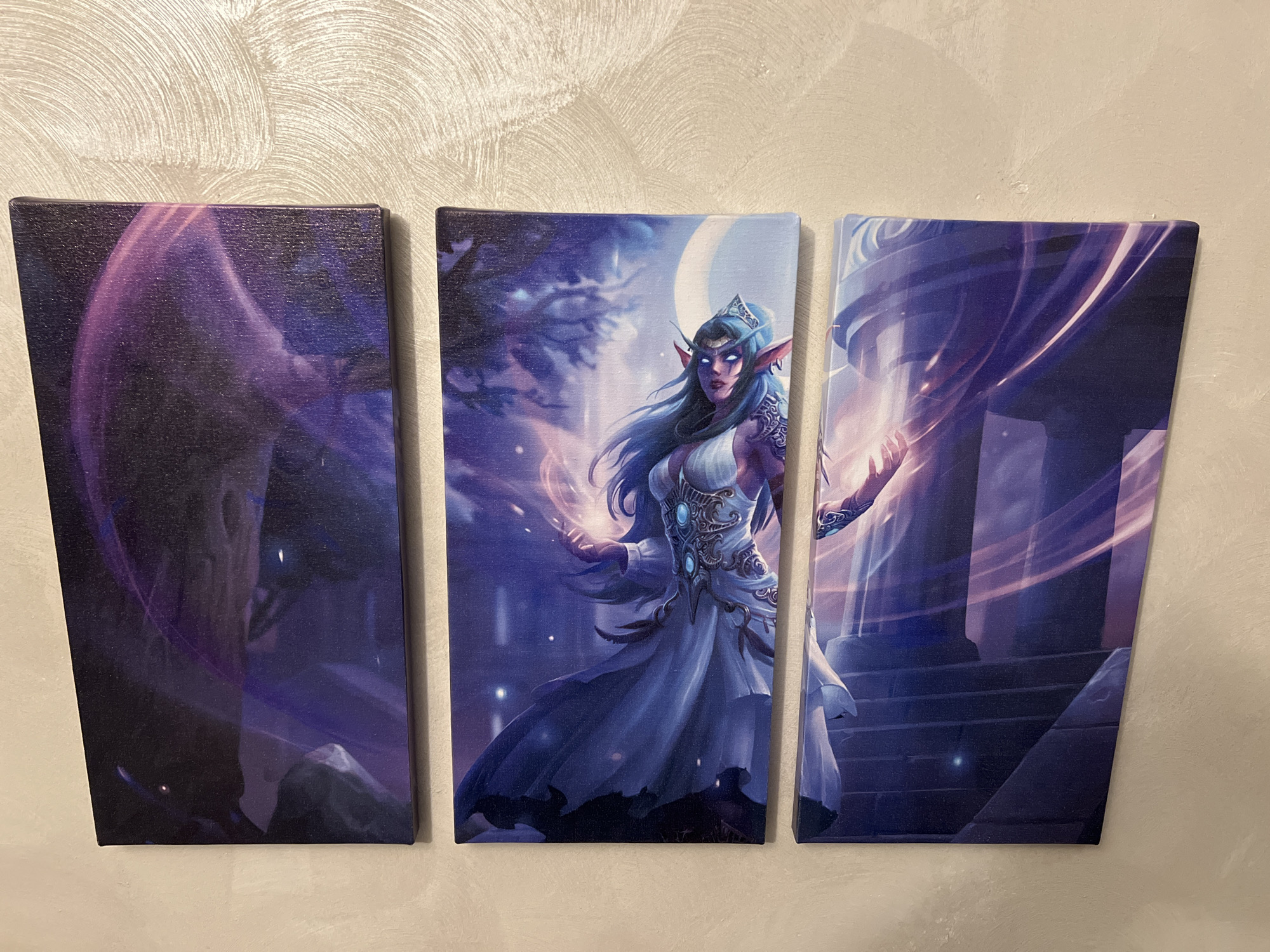 The image shows a triptych hanging on a wall. The artwork depicts an elven woman in a flowing white dress, surrounded by magical energies. The background features elements of fantasy architecture and a night sky with a moon. The leftmost panel focuses on a shadowy structure, while the rightmost panel highlights a columned building with glowing effects. The wall behind the artwork is light beige with a textured pattern. The image was taken indoors. The image is not in Sofia, Bulgaria.