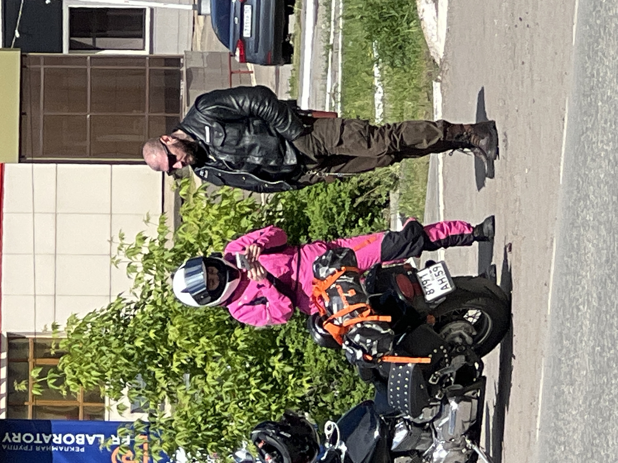 The image shows two people and a motorcycle parked on a sidewalk in Chaikovsky, Russia. A man with a shaved head is standing in a black leather jacket and brown pants. A person in a pink jumpsuit and white helmet is standing next to a motorcycle. The license plate number of the motorcycle is 8191 AH 59. A building is visible in the background, and a blue sign with the words "Laboratory" is also visible. A car is parked in the street in the background.
