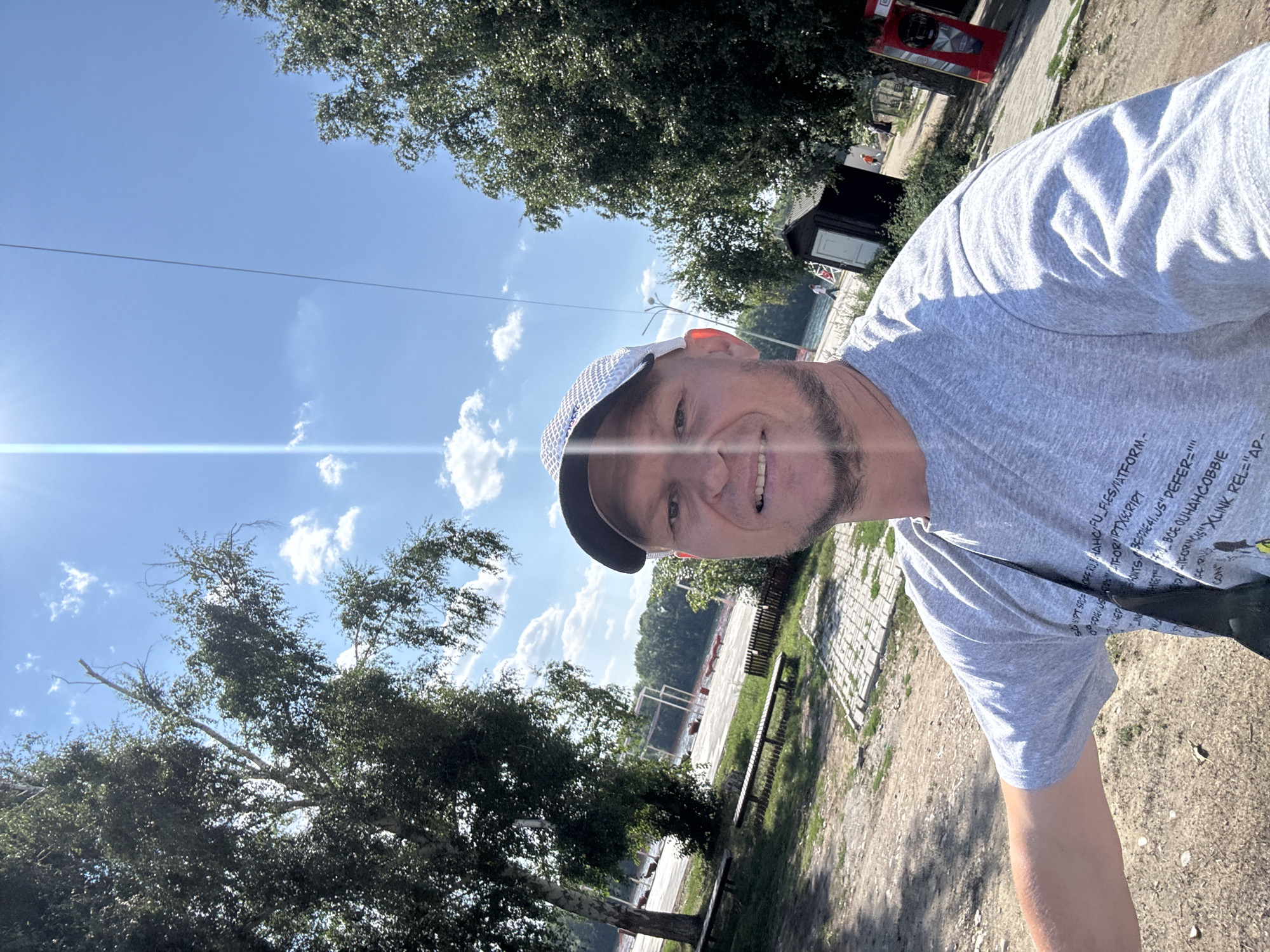 A man with a beard and wearing a grey t-shirt and a baseball cap is taking a selfie outdoors. He is smiling and looking at the camera. The background shows a body of water with trees and some structures on the shore. The sky is blue with some clouds, and the sun is shining brightly. The man appears to be at a recreational area near a lake or river. This location could be the embankment of the Irtysh River in Oskemen, Kazakhstan, which is a popular spot for outdoor activities and relaxation. There are benches and a small wooden building visible in the background, as well as a red structure resembling a phone booth.