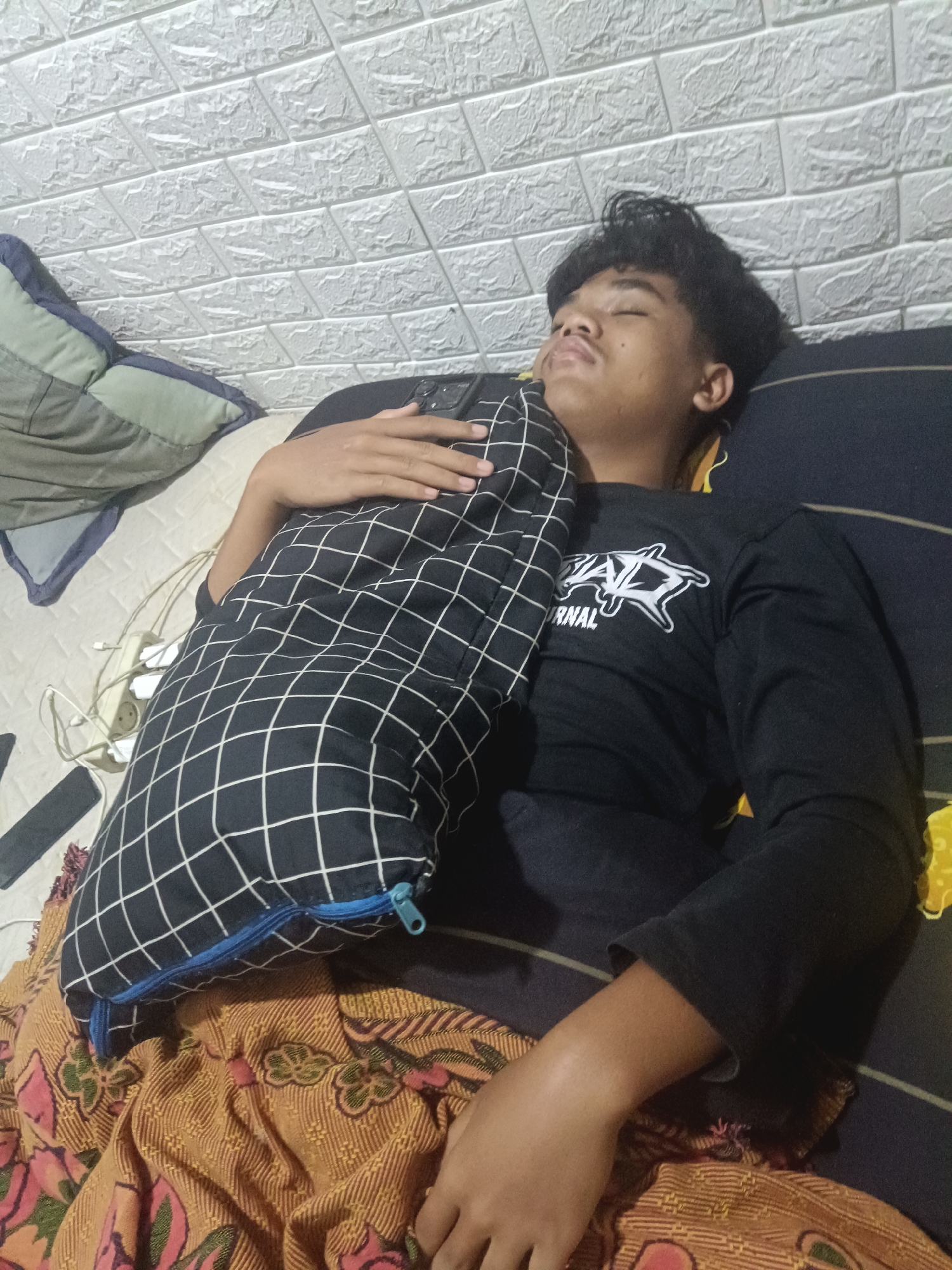 A young person is asleep on a bed, hugging a checkered pillow. They are wearing a black long-sleeved shirt with a white graphic. The bed is covered with a patterned blanket. In the background, a white textured wall is visible. An electrical outlet strip with several plugs is also present on the bed. The scene appears to be indoors, likely in a residential setting in Klepu, Indonesia.
