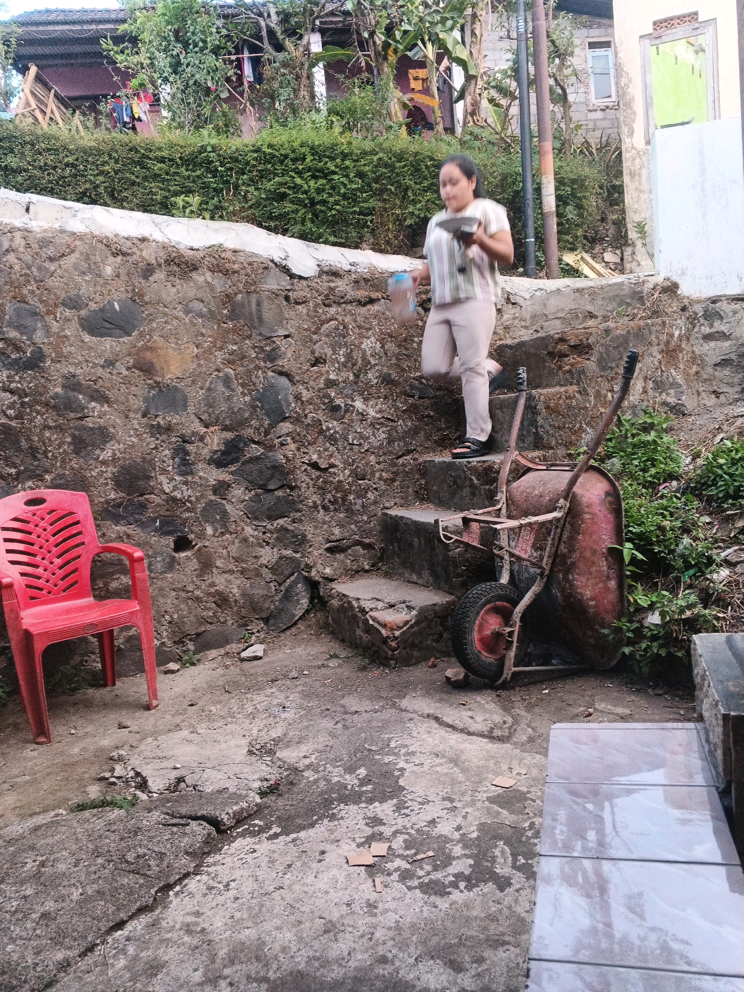 In Keji, Indonesia, a woman walks down a set of outdoor stairs carrying a bottle of water and a tool. She is wearing a striped shirt and light-colored pants, and appears to be moving from left to right. Beside the stairs, a red plastic chair sits empty next to a rough stone wall. A rusty wheelbarrow with a red bucket rests on the landing at the bottom of the stairs, surrounded by green foliage and debris. The background reveals a hillside with buildings and lush vegetation, suggesting a semi-rural or residential setting. The ground is a mix of cracked concrete and dirt.